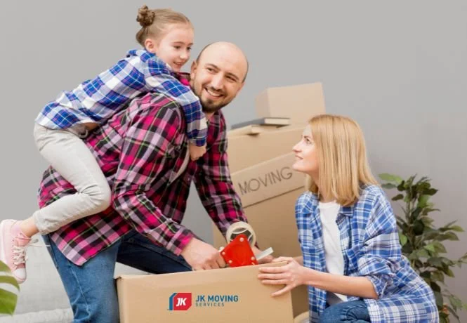 A family of three, with a father carrying a young girl on his back, packing and preparing to move in a room with moving boxes, including one labeled 'MOVING', and a woman sitting on the floor.