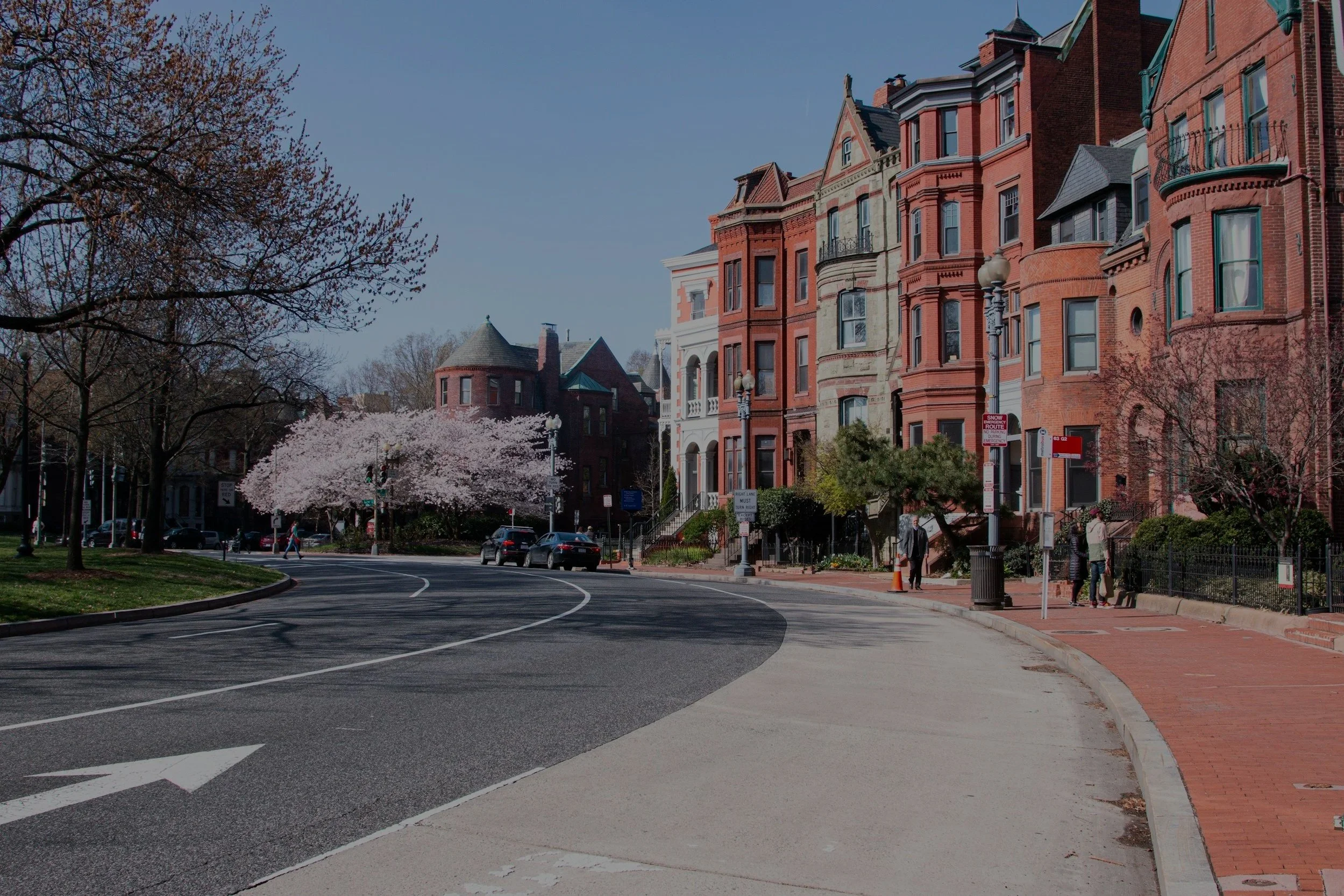 A city street with Victorian-style brick buildings, a blossoming cherry tree, pedestrians, and parked cars on a sunny day.
