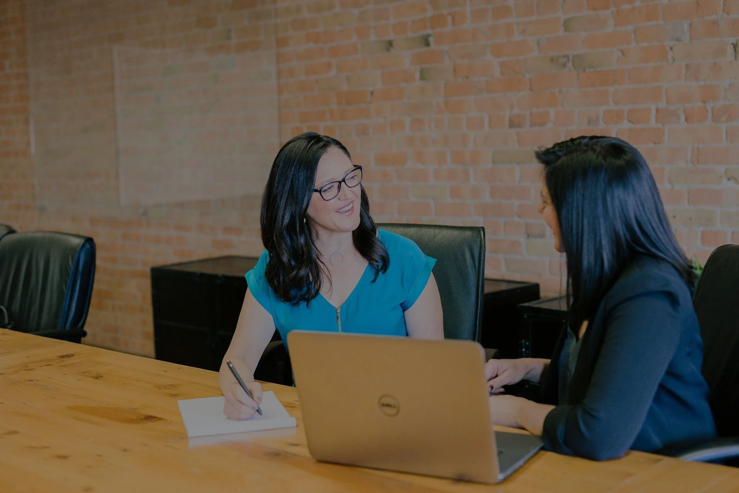 Two women having a business meeting in a conference room with a brick wall background, one woman with black hair and glasses, holding a pen and notebook, smiling at the other woman with dark hair, working on a laptop.