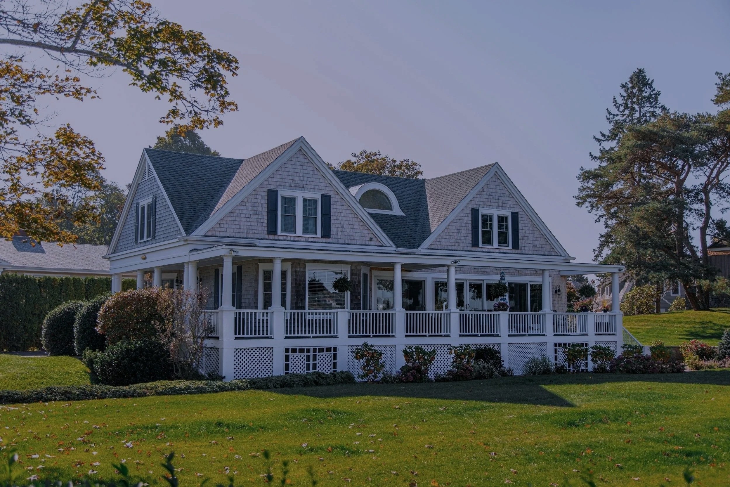 A large, white house with a wrap-around porch, multiple windows, gabled roof, surrounded by green lawn and trees.