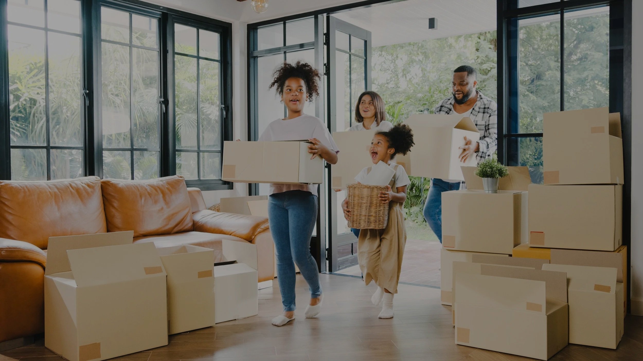 A family is moving into a new home, carrying cardboard boxes. The room has large windows and a brown leather sofa, with boxes stacked around.
