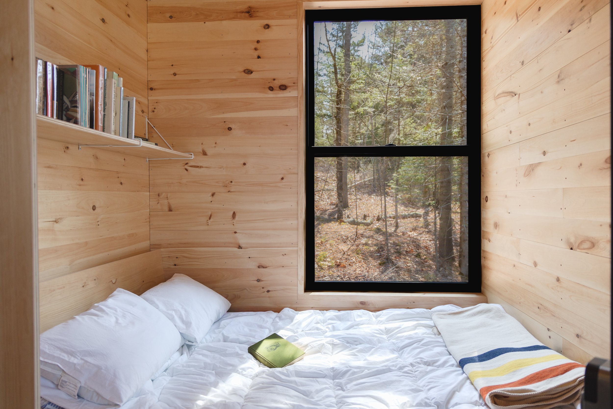 A cozy bedroom with wooden walls and a large window showing a forested outdoor scene. The bed has white bedding, a striped towel, and a closed book on it.