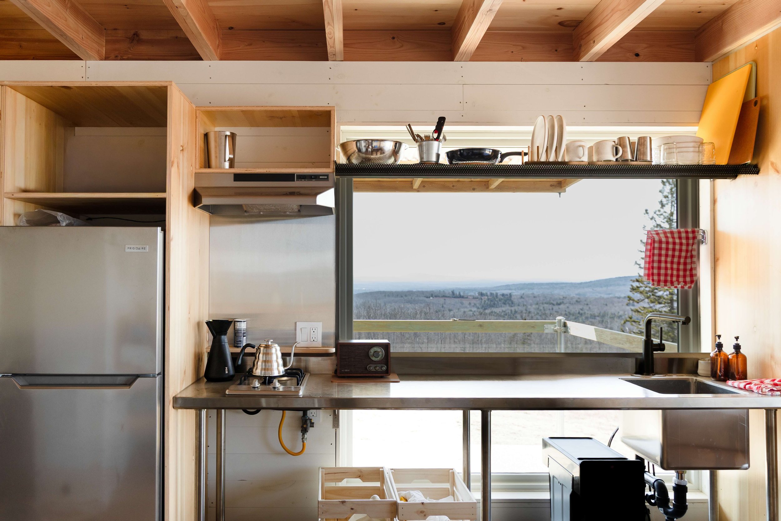 A kitchen with wooden cabinets and shelves, a small refrigerator, and a large window showing a scenic outdoor landscape with trees and a cloudy sky. There are kitchen utensils, dishes, and appliances visible on the counter and shelves.