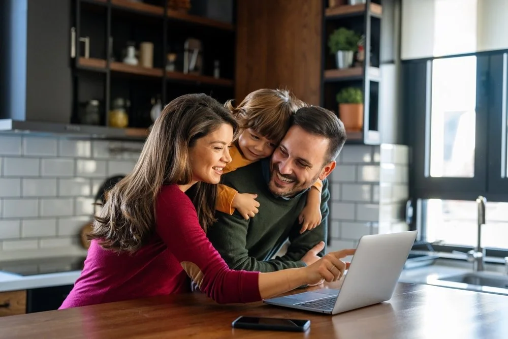 Happy family of three using a laptop in a modern kitchen, with the mother pointing at the screen.