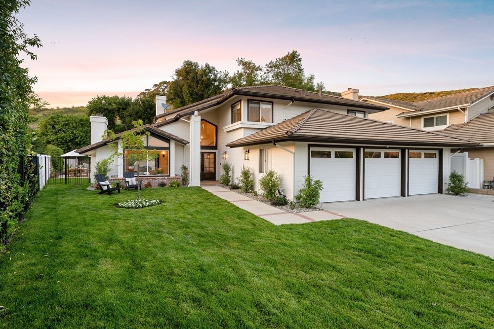 A modern two-story house with a white exterior, brown roof, and three-car garage, surrounded by a lush green lawn with small shrubs and a tree near the entrance, under a pastel sky at sunset.