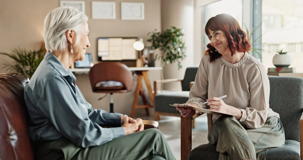 A woman with gray hair and earrings talks to a younger woman with dark red hair, holding a notepad and pen, in a cozy, well-lit room with plants and framed pictures.