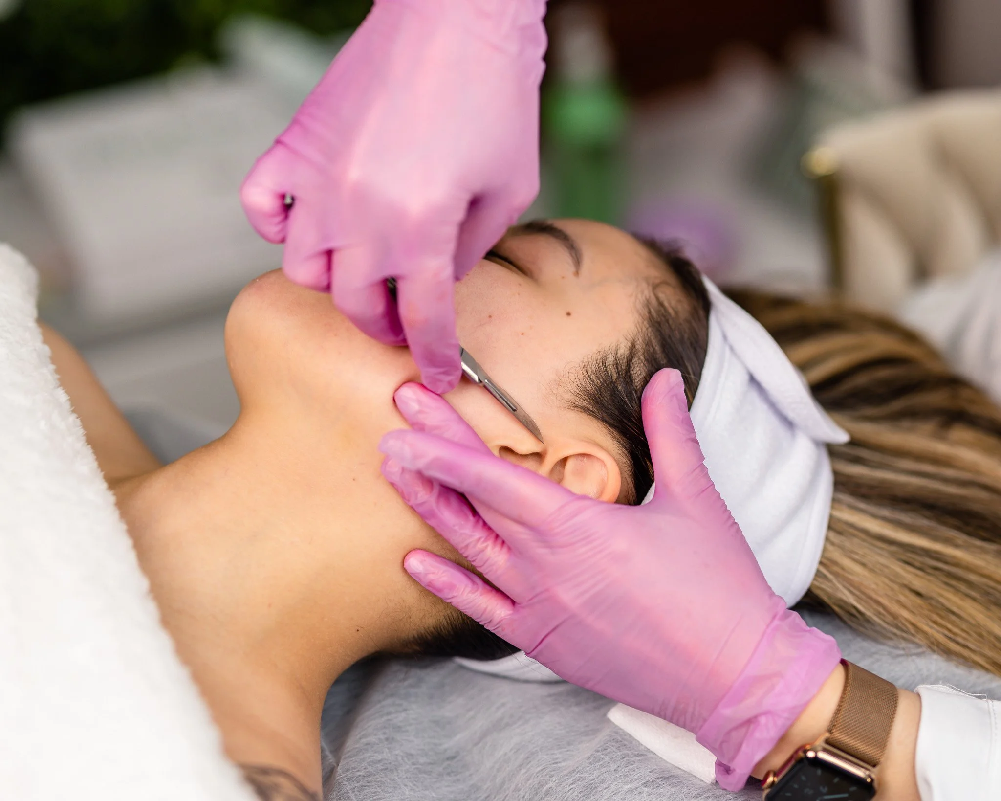 A woman receiving a beauty treatment or facial, lying down with a headband, while a professional wearing pink gloves uses a tool near her face.