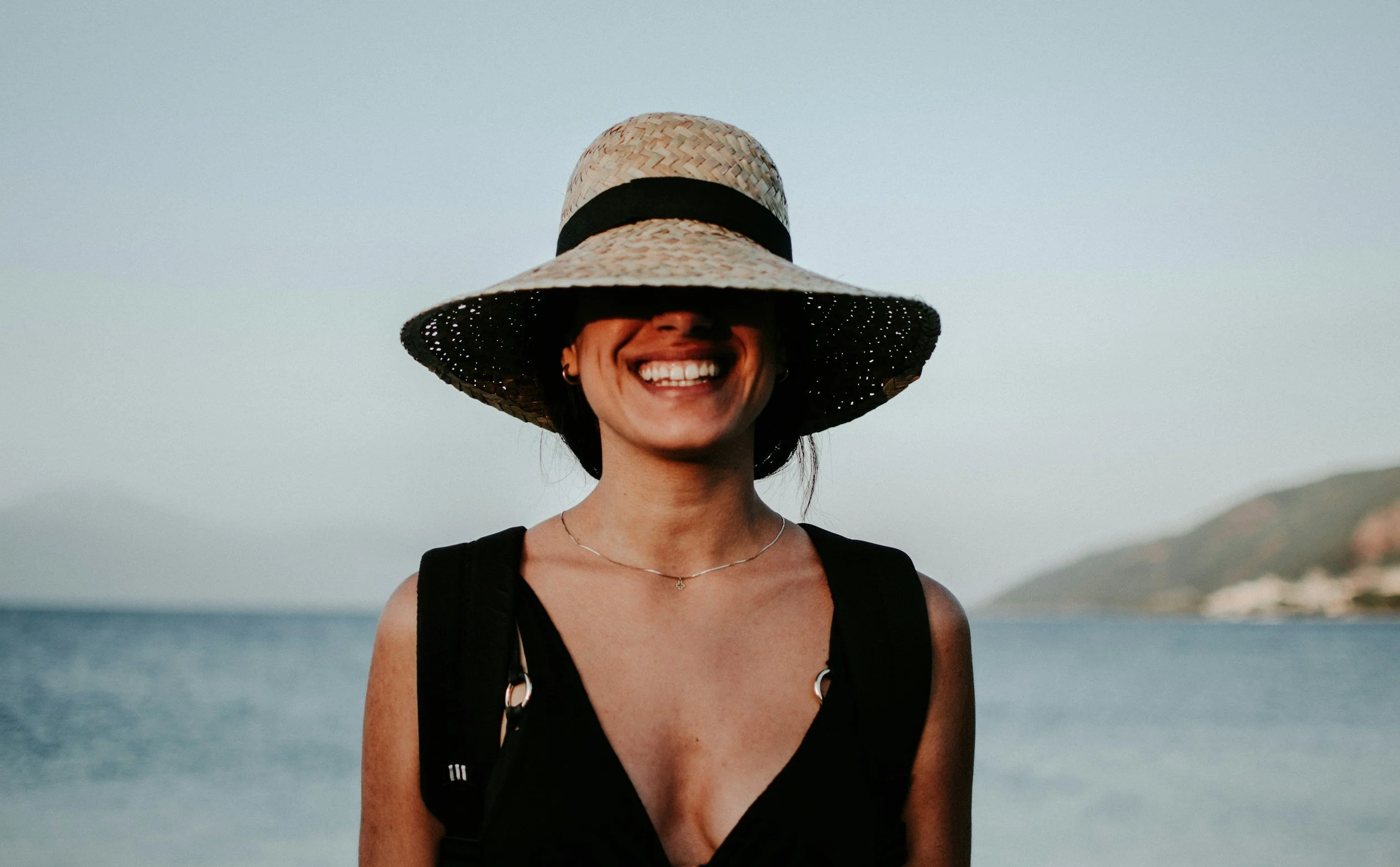 Smiling woman wearing a wide-brimmed straw hat with black band, standing near water with hills in the background.