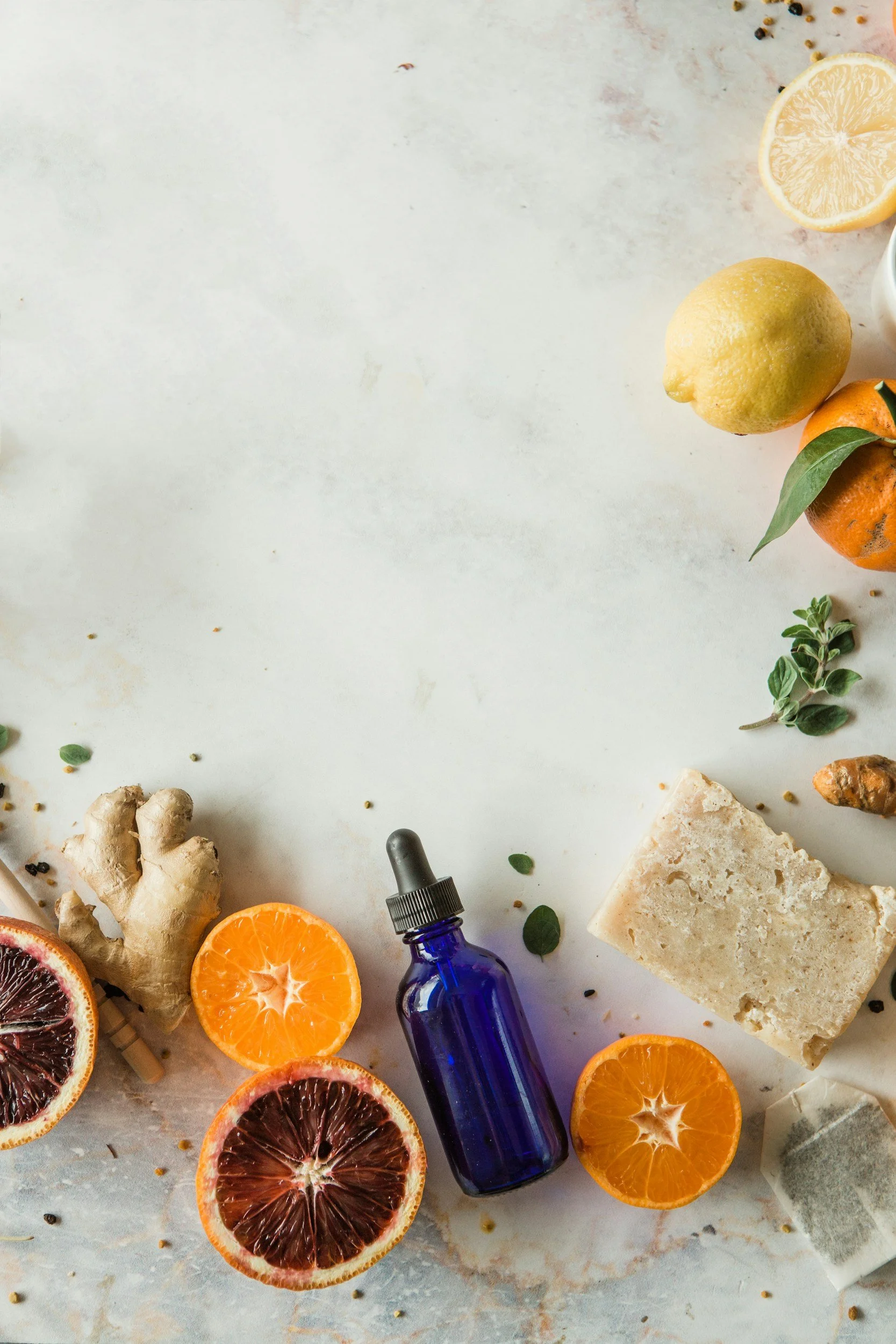 Flat lay of natural remedies and ingredients, including blood oranges, lemon, ginger root, a blue dropper bottle, soap, and herbs, arranged on a light surface.