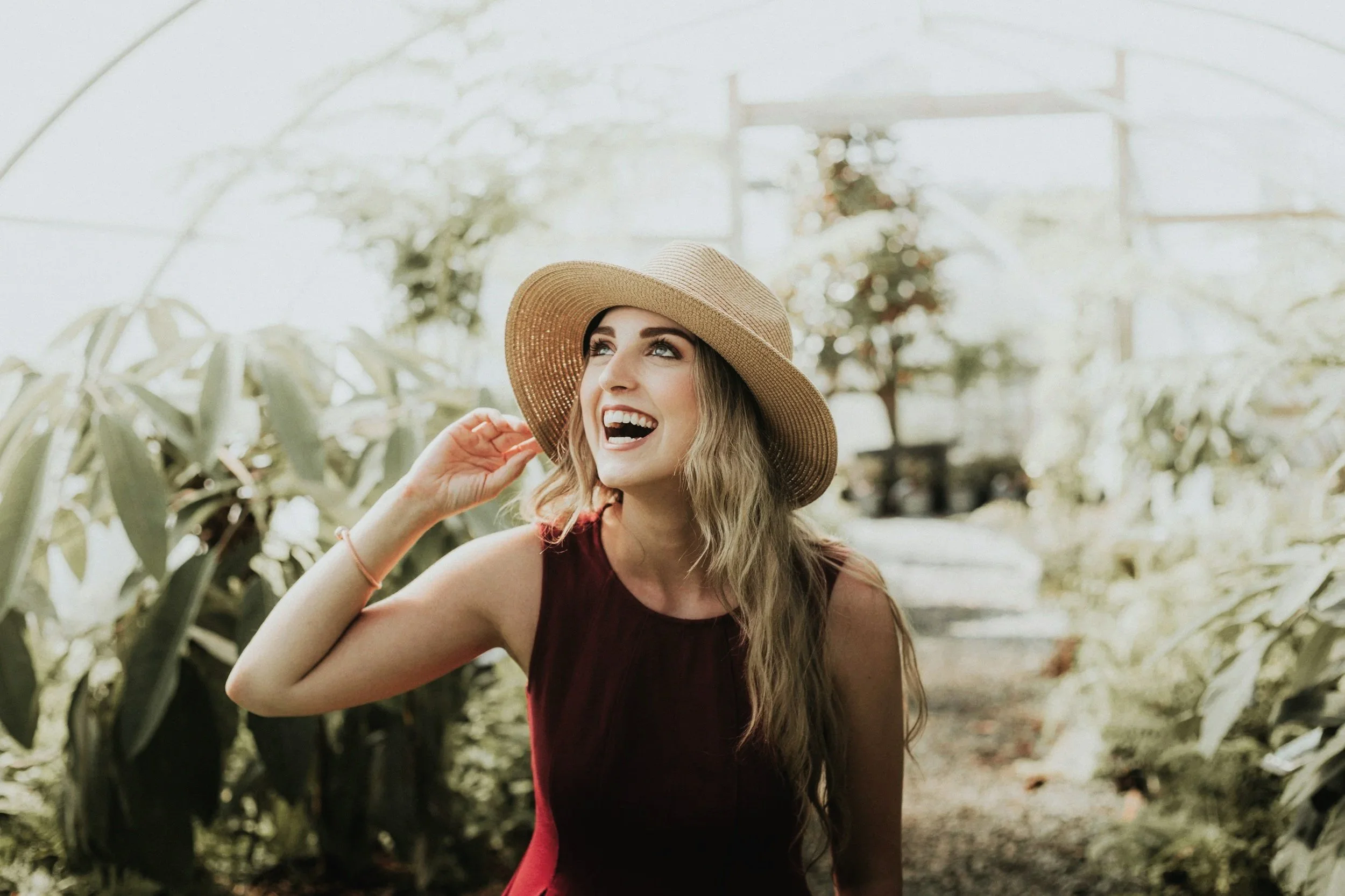 A woman with long wavy hair smiling and wearing a large straw hat and sleeveless red dress inside a greenhouse filled with green plants.