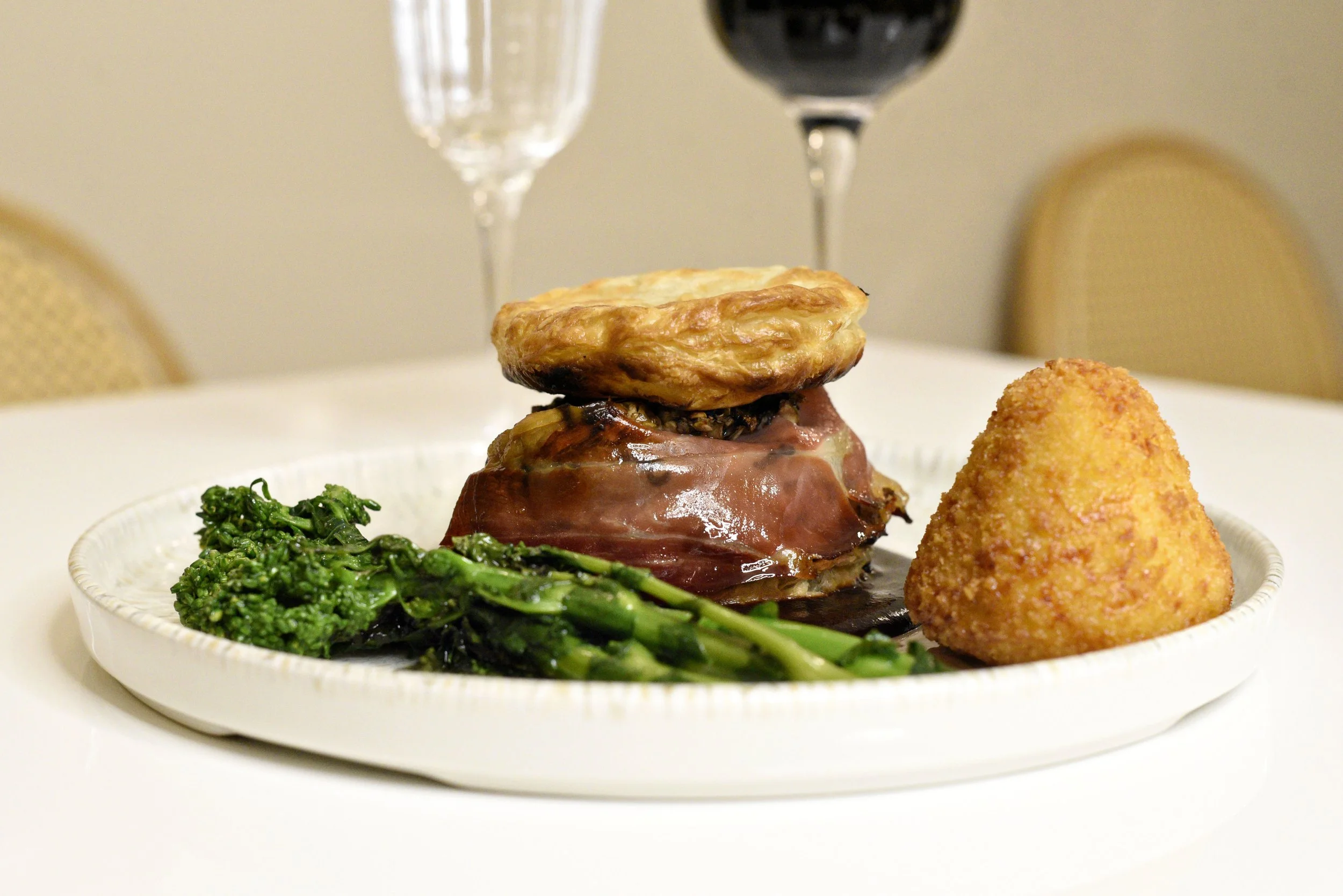 Café Nelo — A plated gourmet dish featuring a layered savory pastry on top of a slice of cured meat, accompanied by cooked greens and a crispy, golden croquette. In the background, two glasses of wine, one with white wine and one with red wine.