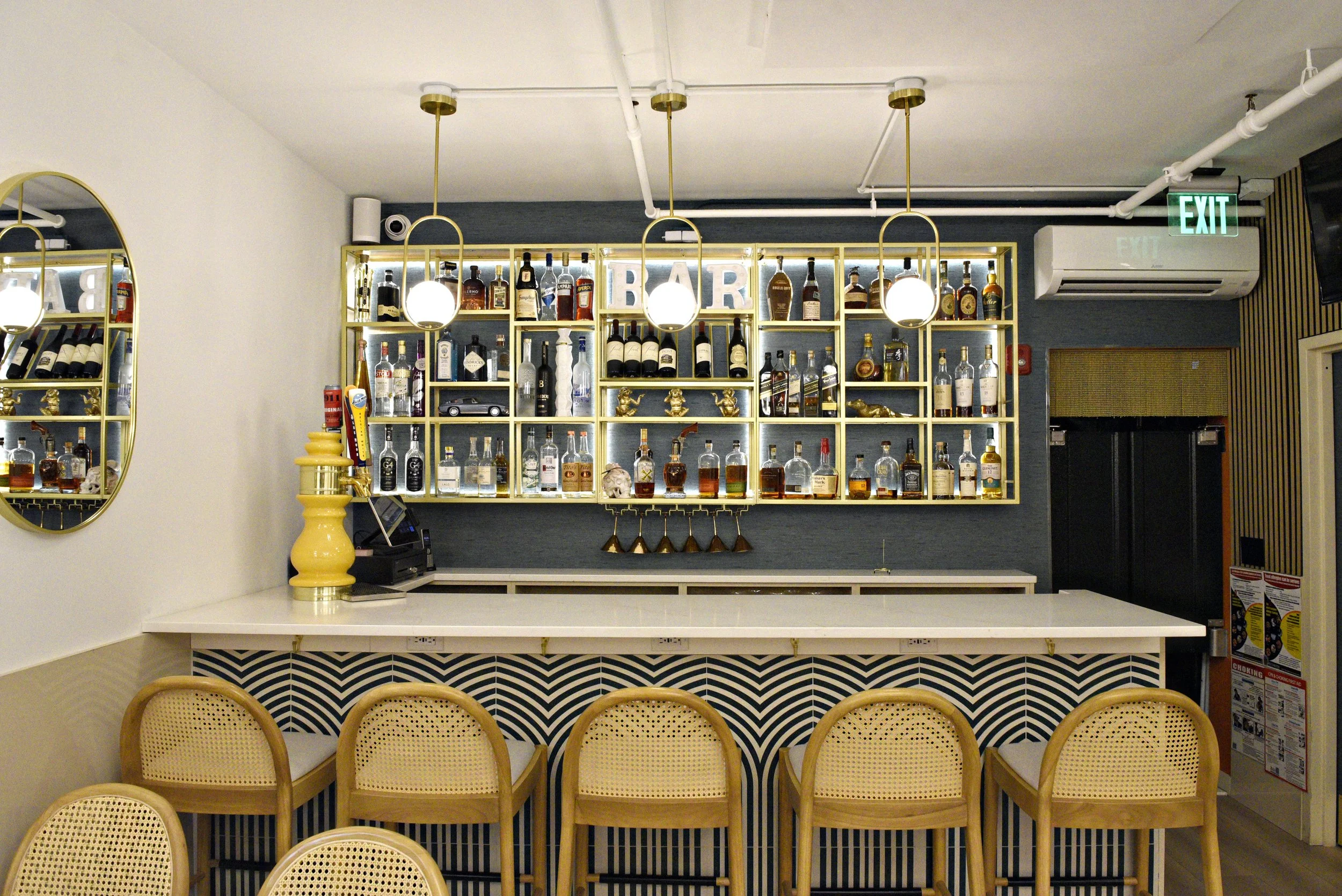Café Nelo — A bar with shelves of assorted liquor bottles and a sign that says 'BAR' in the background. There are pendant lights hanging from the ceiling and seating with woven rattan chairs with cushioned seats in the foreground.