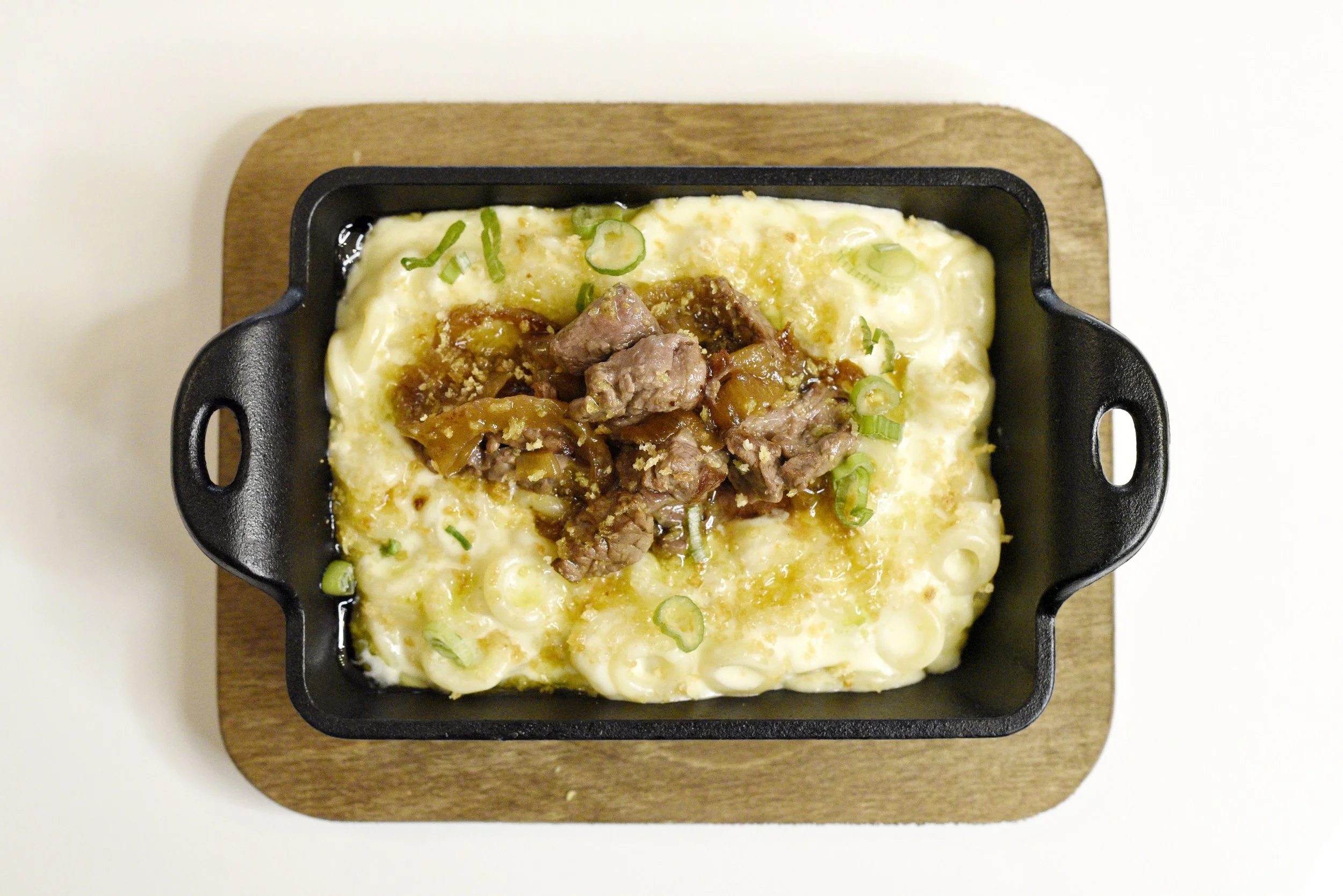 Café Nelo — Baked macaroni and cheese with a serving of beef and vegetable stew on top, garnished with chopped green onions, in a small black cast iron skillet on a wooden board.