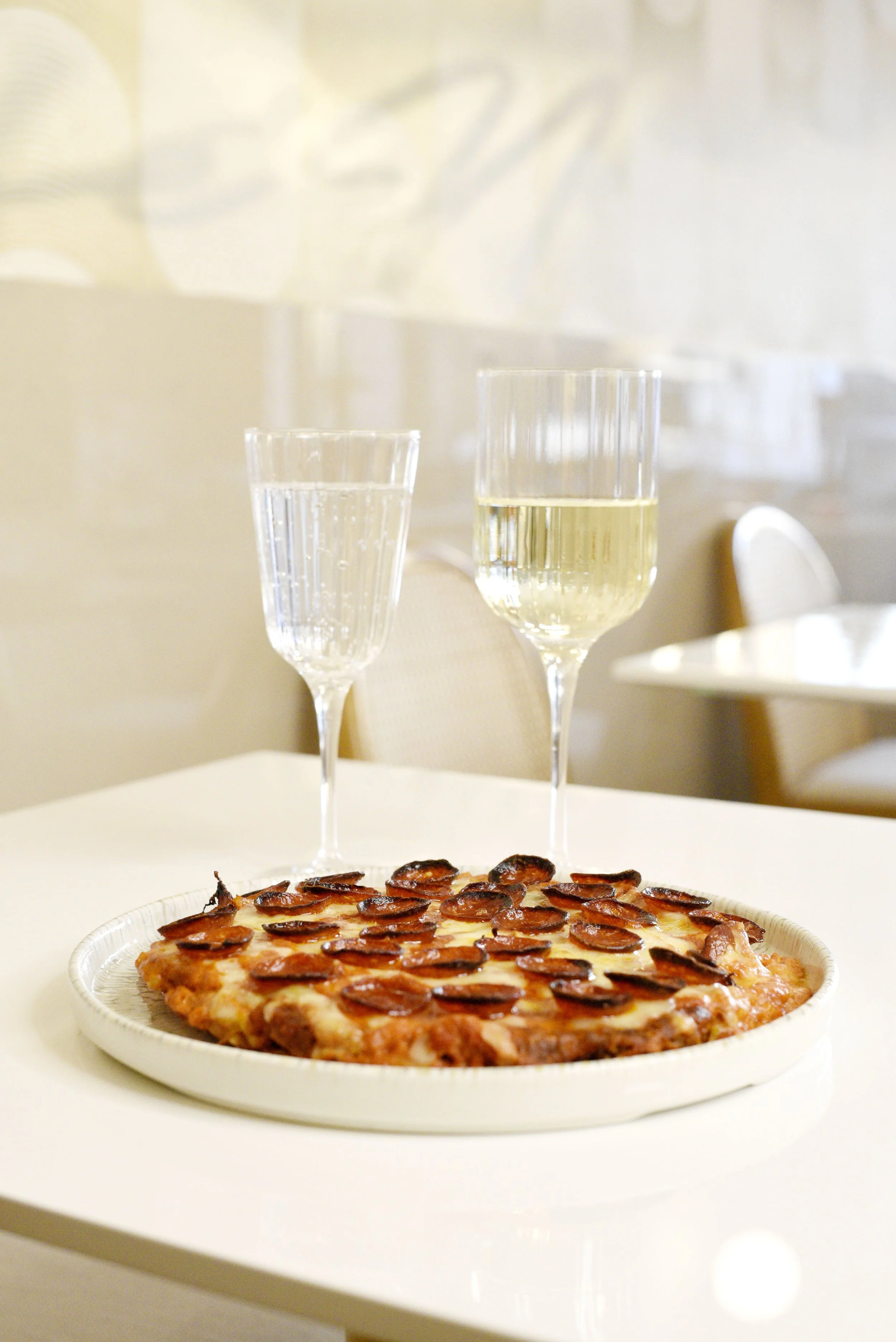 Café Nelo — Pizza with pepperoni slices on a white table, two glasses of white wine, and a blurred background with chairs and wall art.