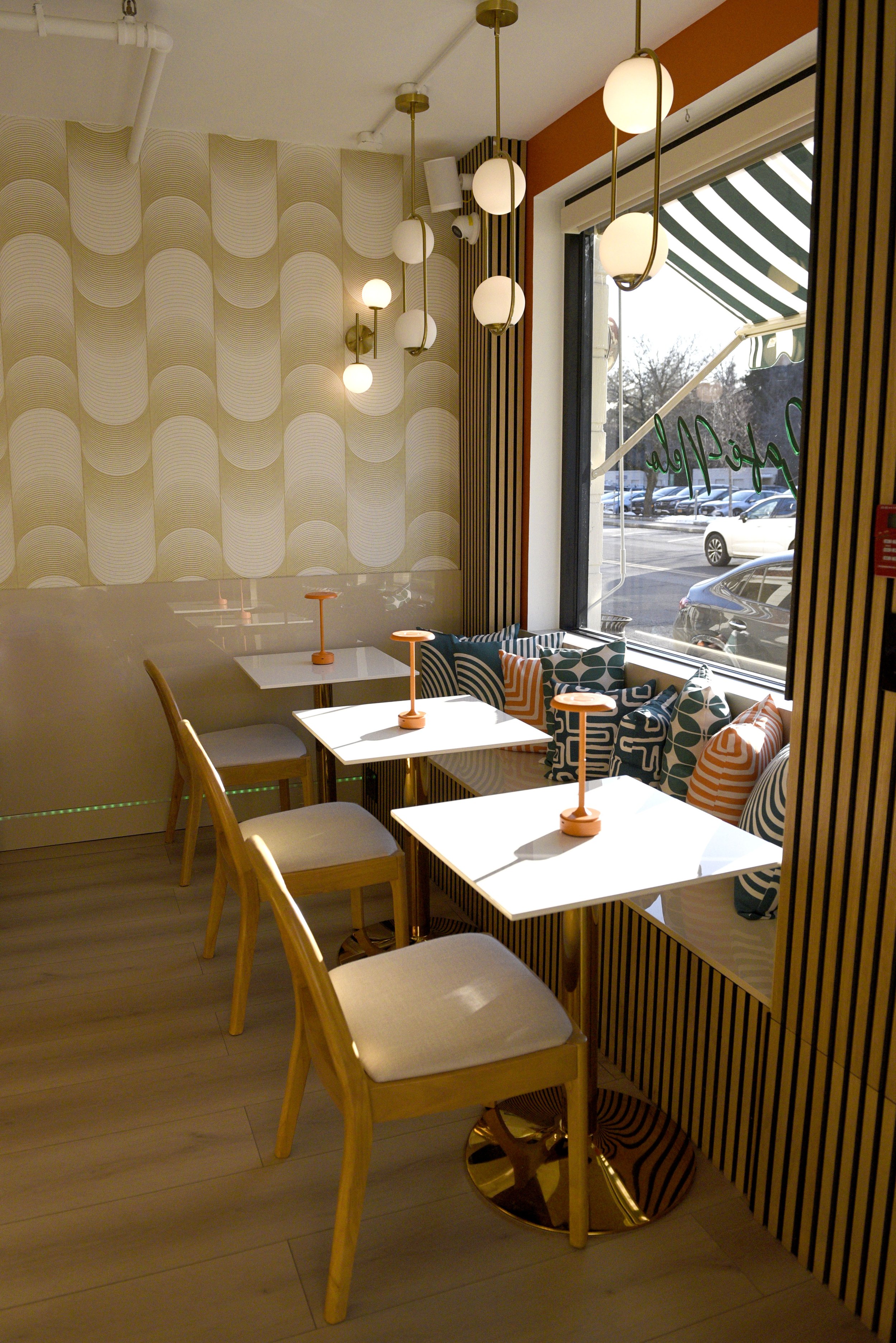 Café Nelo — Interior of a modern cafe or restaurant with wooden chairs, white tables, patterned cushions, and decorative lighting fixtures with globes hanging from the ceiling. There is a window showing cars outside and a striped awning.