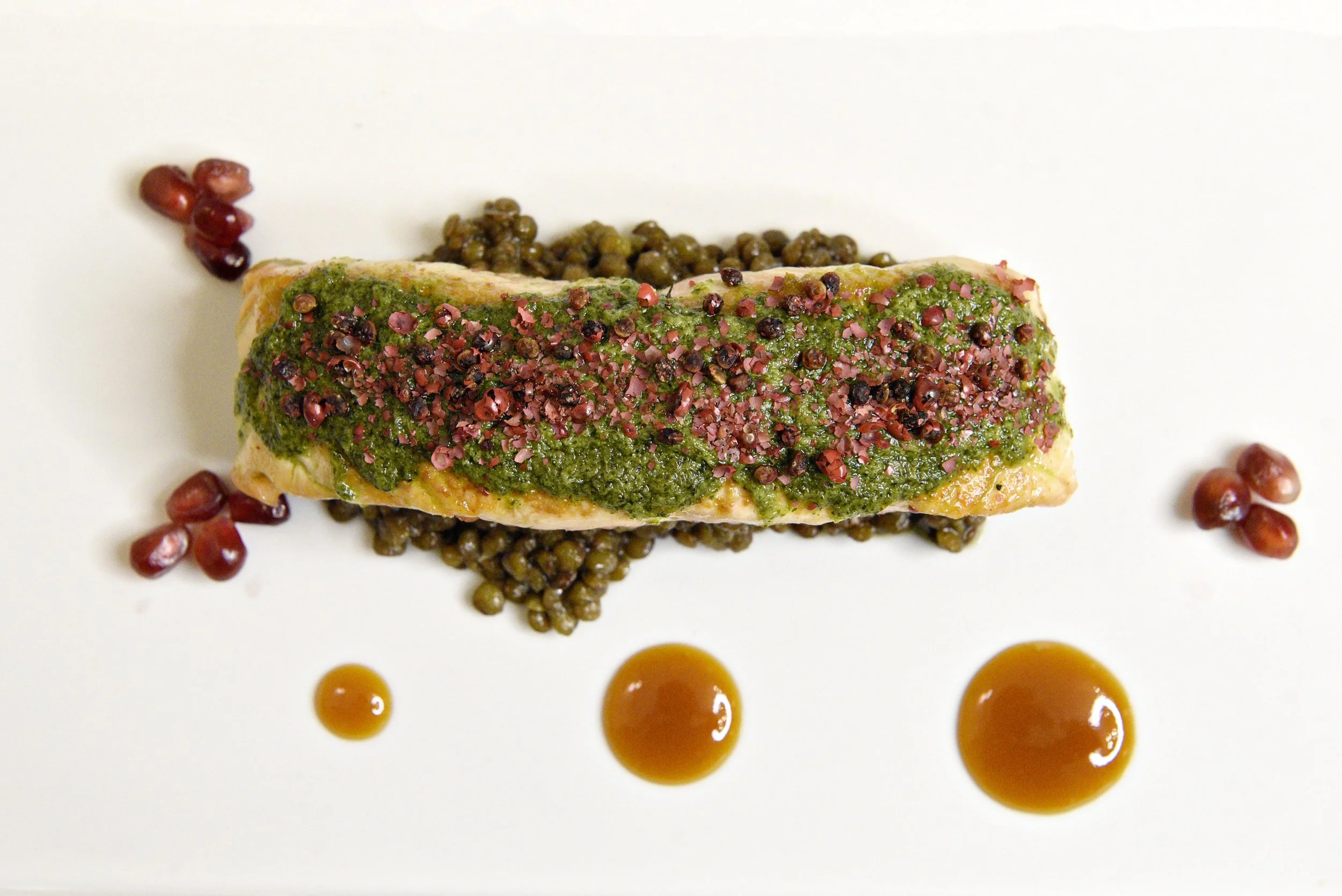 Café Nelo — A plated gourmet dish featuring a piece of fish topped with green pesto and pink peppercorns, surrounded by scattered pomegranate seeds and small droplets of orange-colored sauce on a white plate.