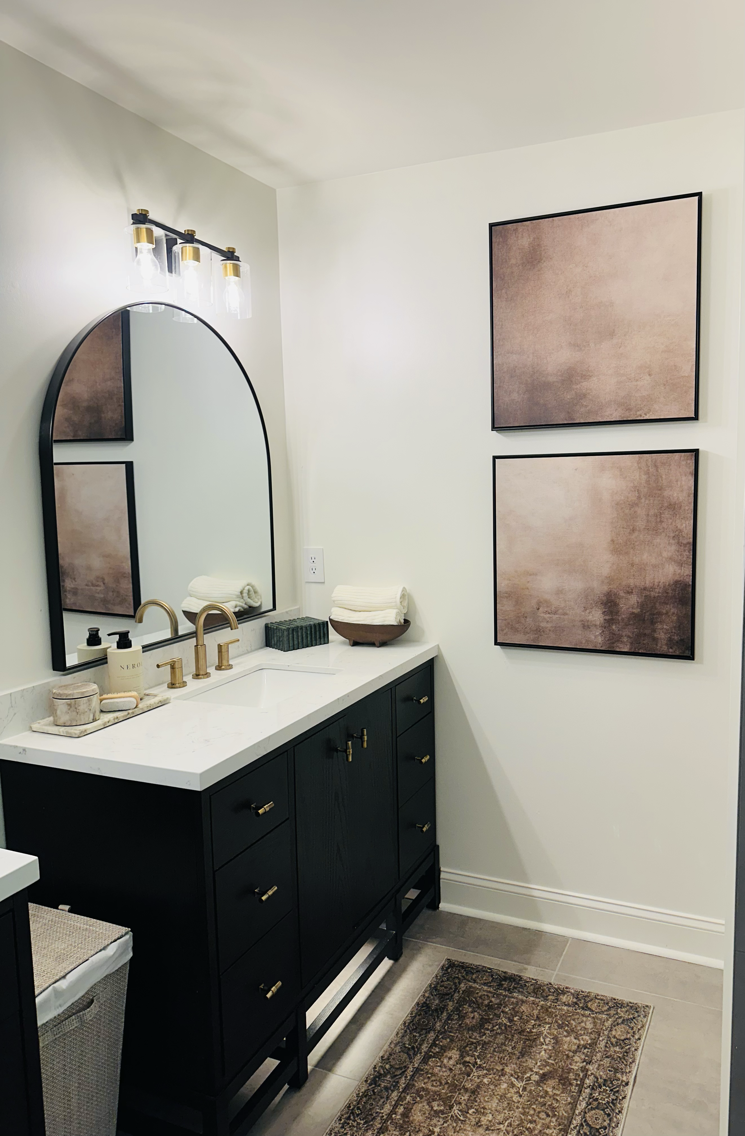 Modern bathroom vanity with a black cabinet, white countertop, gold fixtures, and a large mirror. Decor includes rolled towels, soap, a small basket, and two framed art pieces on the wall.