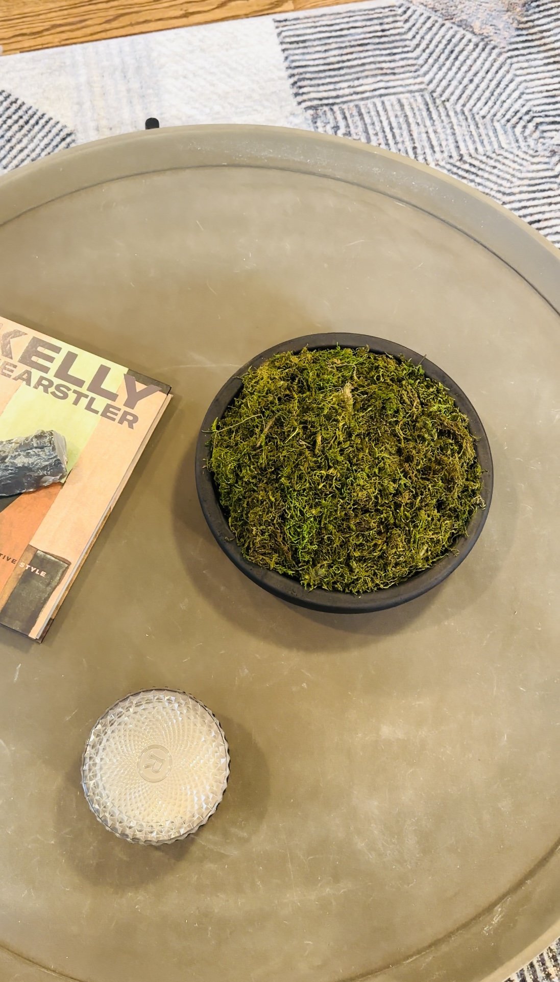 A round black bowl filled with green moss placed on a beige table. To the left, there's a magazine titled 'KELLY EARBSTER' and a small textured glass candle holder.