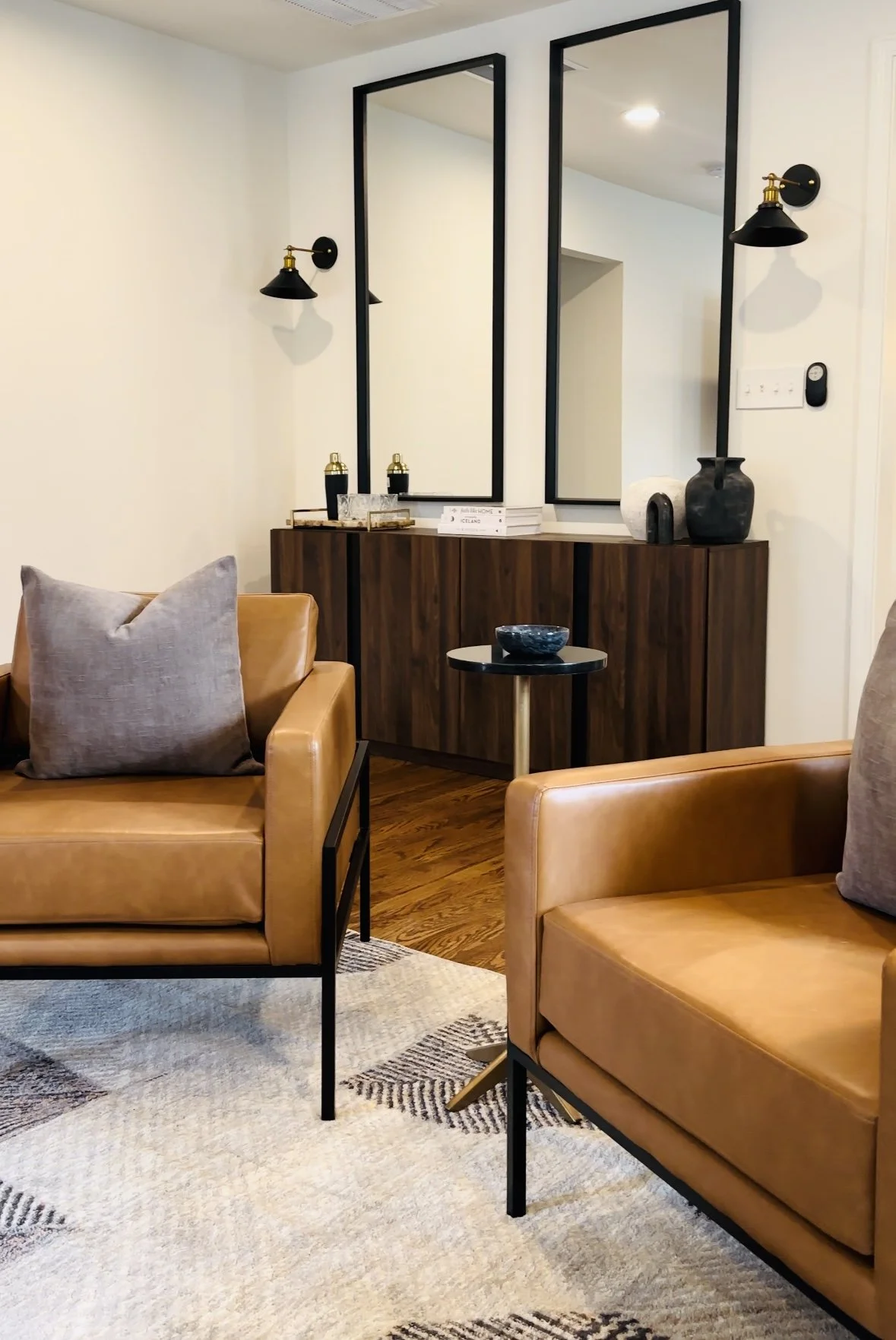 Living room with two tan leather armchairs, a black and gold side table, a wooden sideboard, and two large vertical mirrors on the wall, decorated with black and gold vases and wall sconces.