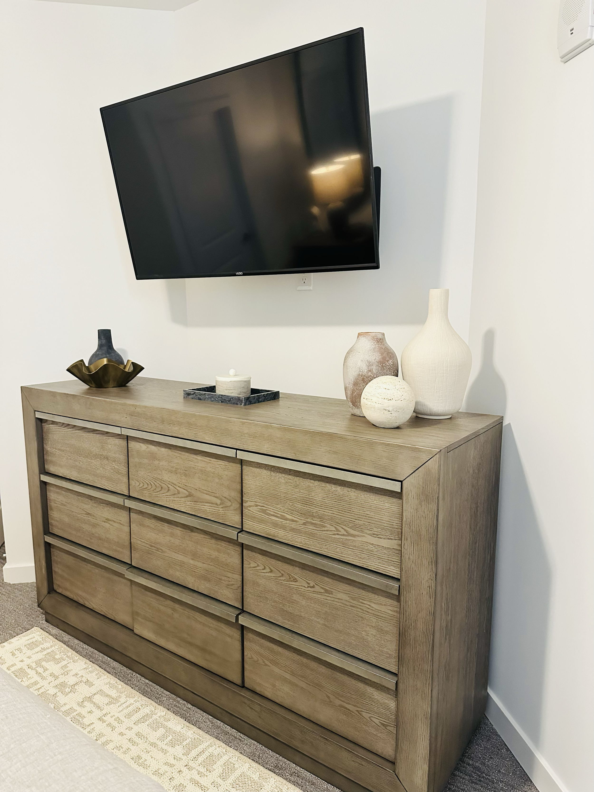 Wooden dresser with decorative vases and tray, mounted flat-screen TV on white wall.