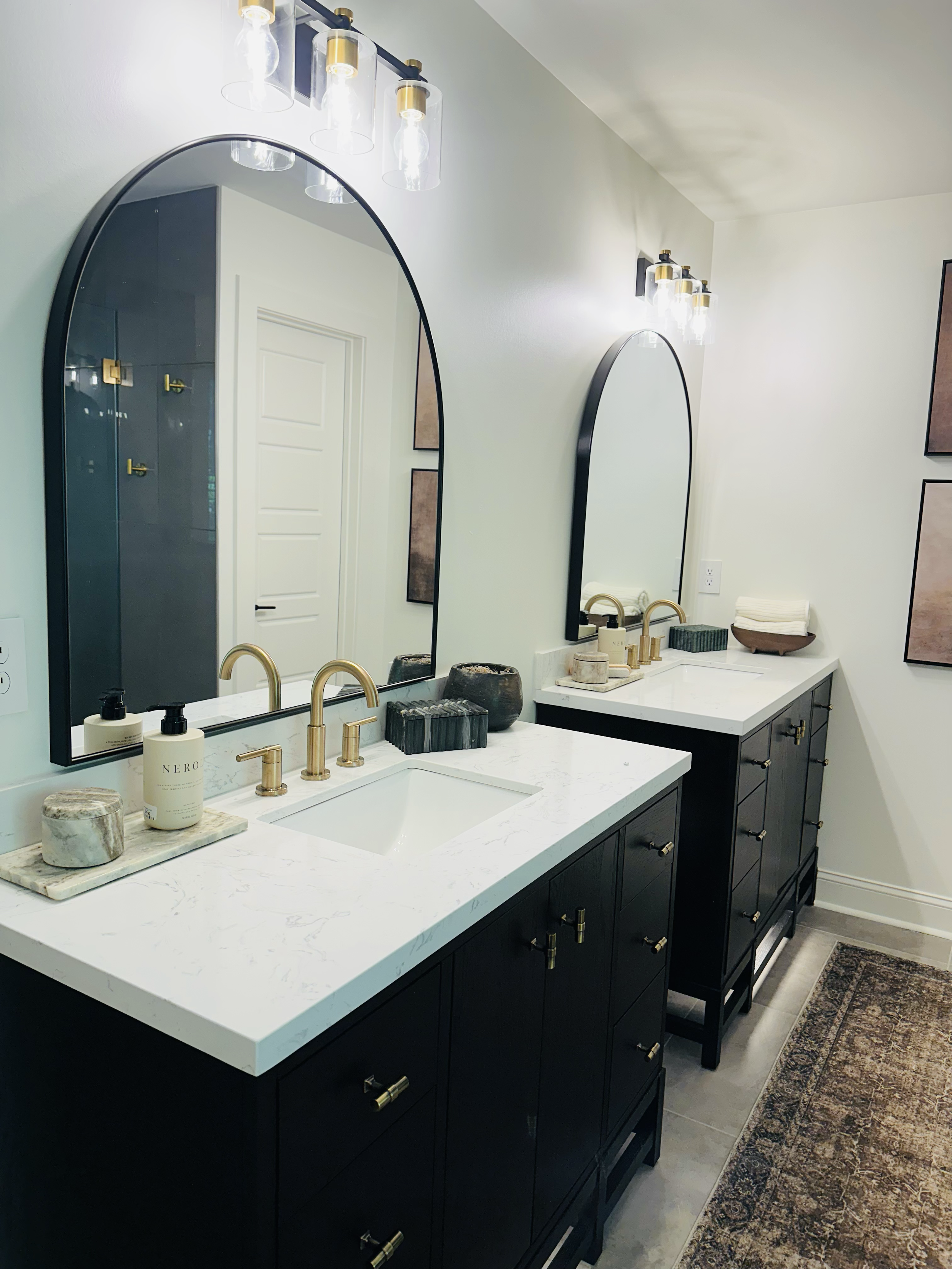 A modern bathroom vanity with double sinks, black cabinets, white marble countertops, gold fixtures, oval mirrors, and decorative items such as soap dispensers, candles, and a basket of towels.