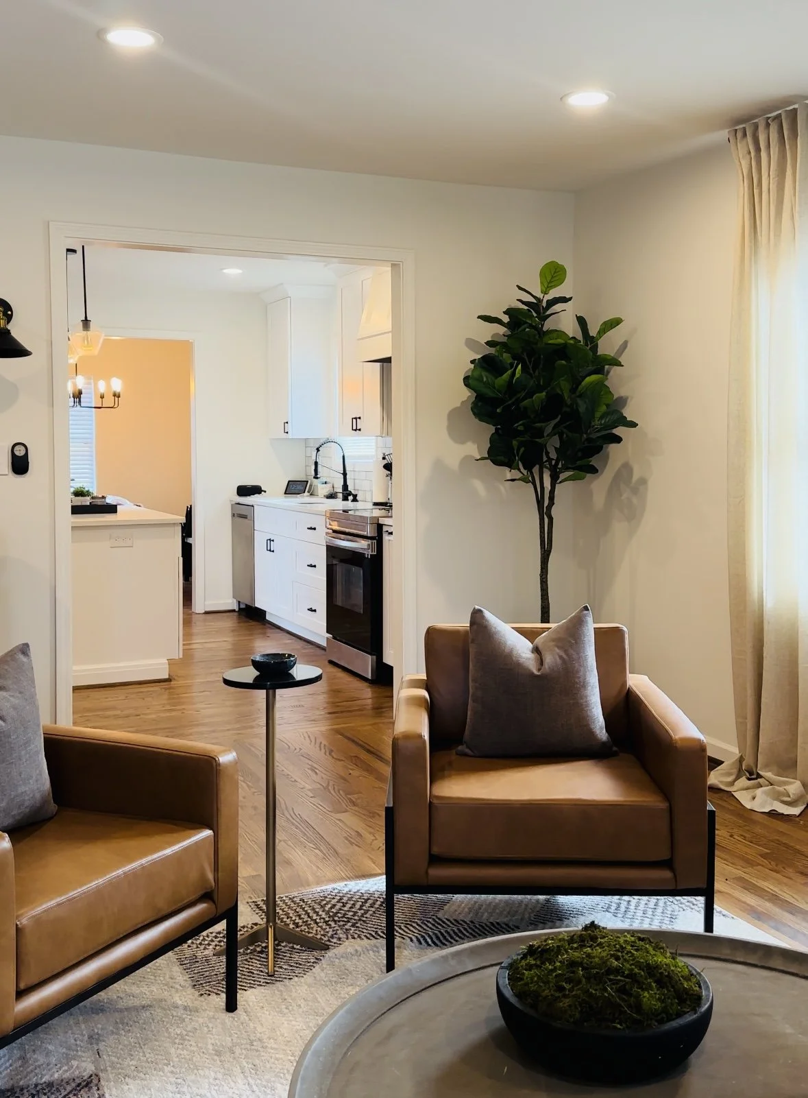 Living room with tan leather armchair, gray pillow, white walls, wooden floor, and a large green potted plant; doorway leads to kitchen with white cabinets and black appliances; window with beige curtains on the right.