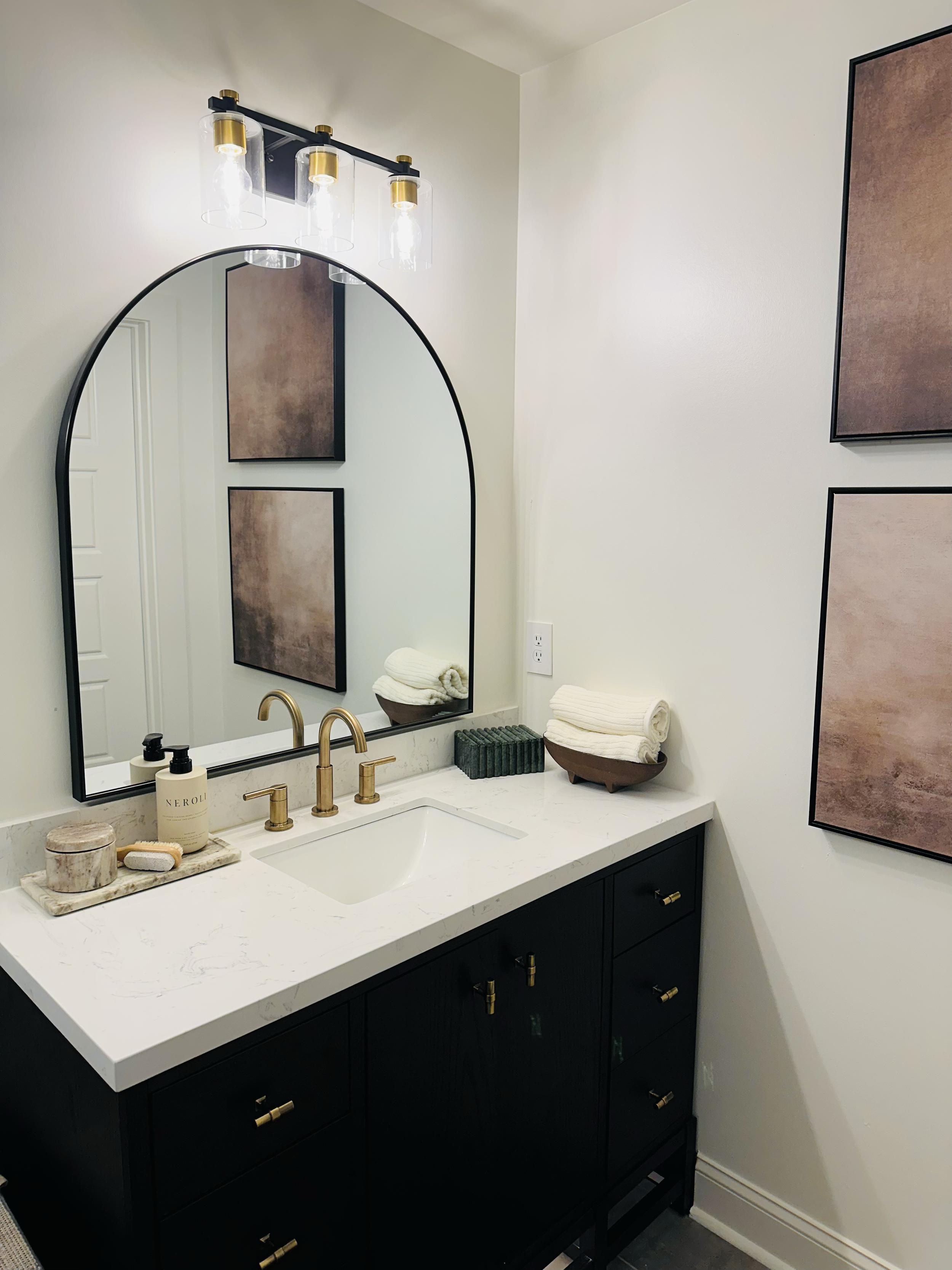 Modern bathroom vanity with a white marble countertop, black cabinetry with gold handles, and a gold faucet. Above the sink is a large oval mirror and a three-light fixture with exposed bulbs and glass covers. Decor includes rolled white towels, a so