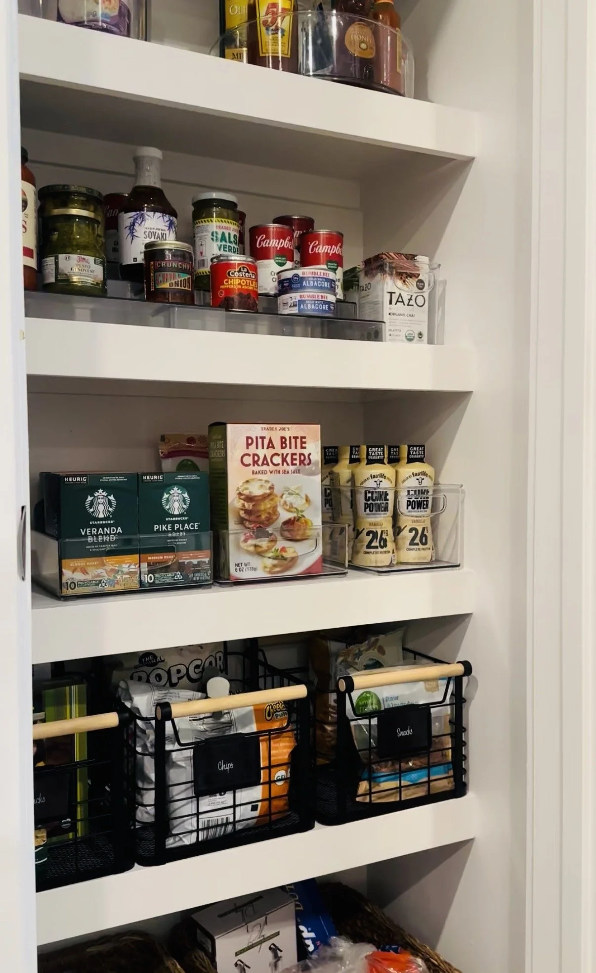 Pantry shelf containing canned goods, boxes of tea and chips, bottles of sauces and condiments, and organized snack baskets.