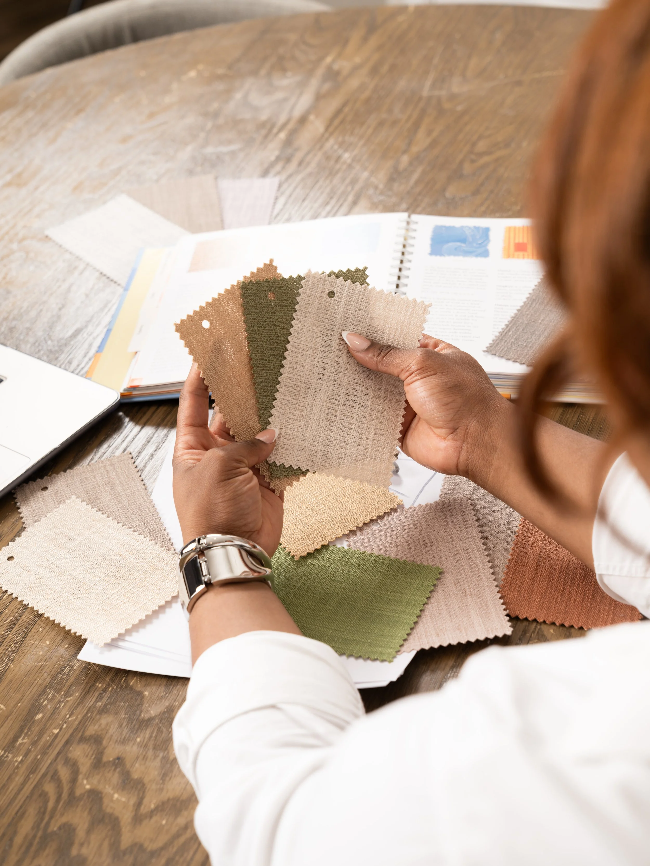 A person examining fabric swatches for interior design or upholstery, with sample book and fabric samples on a wooden table.