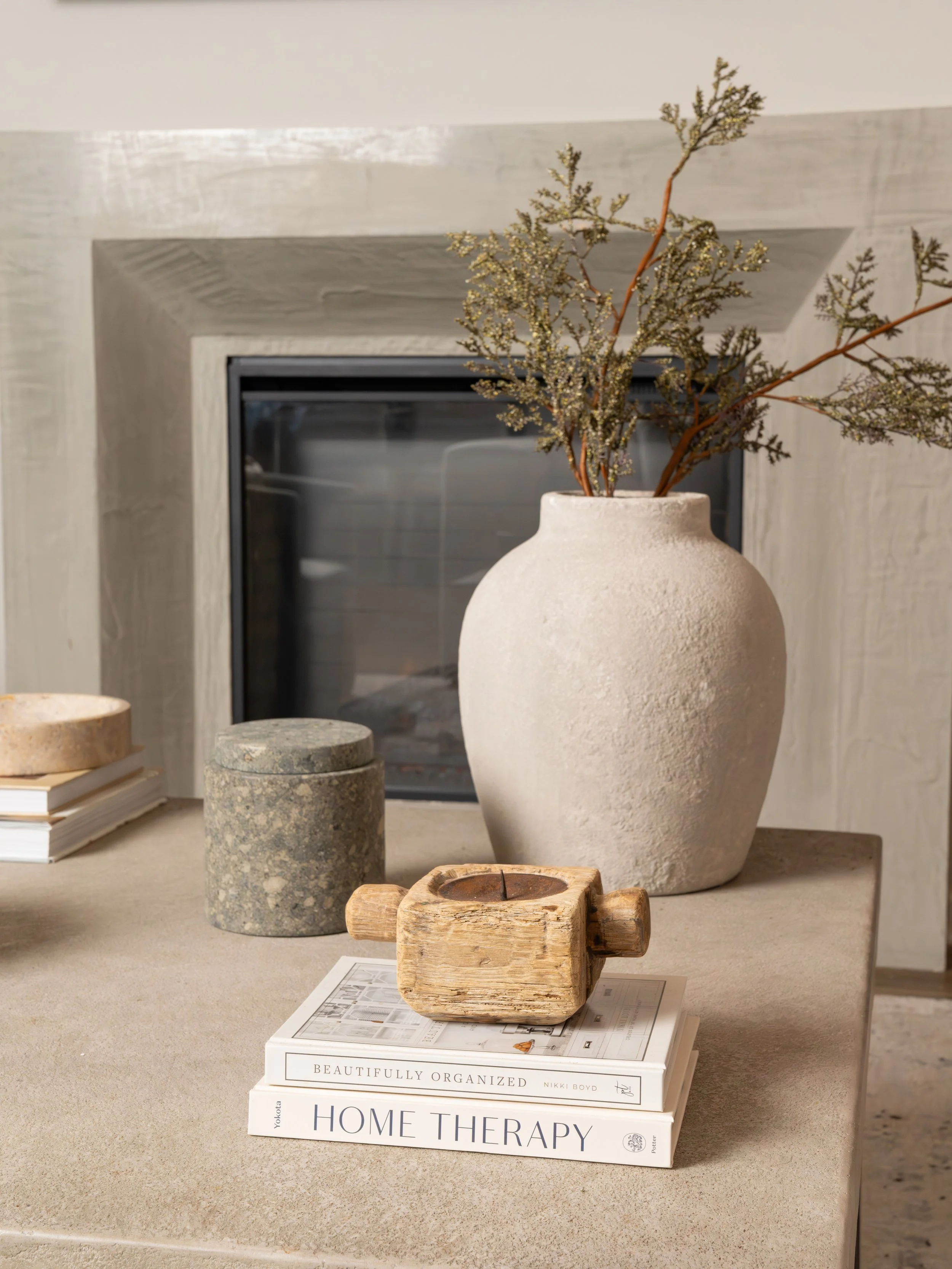 Decorative items on a beige table in front of a fireplace, including a large white vase with dried branches, a gray stone container, a small wooden candle holder, and two stacked books titled 'Beautifuly Organized' and 'Home Therapy'.