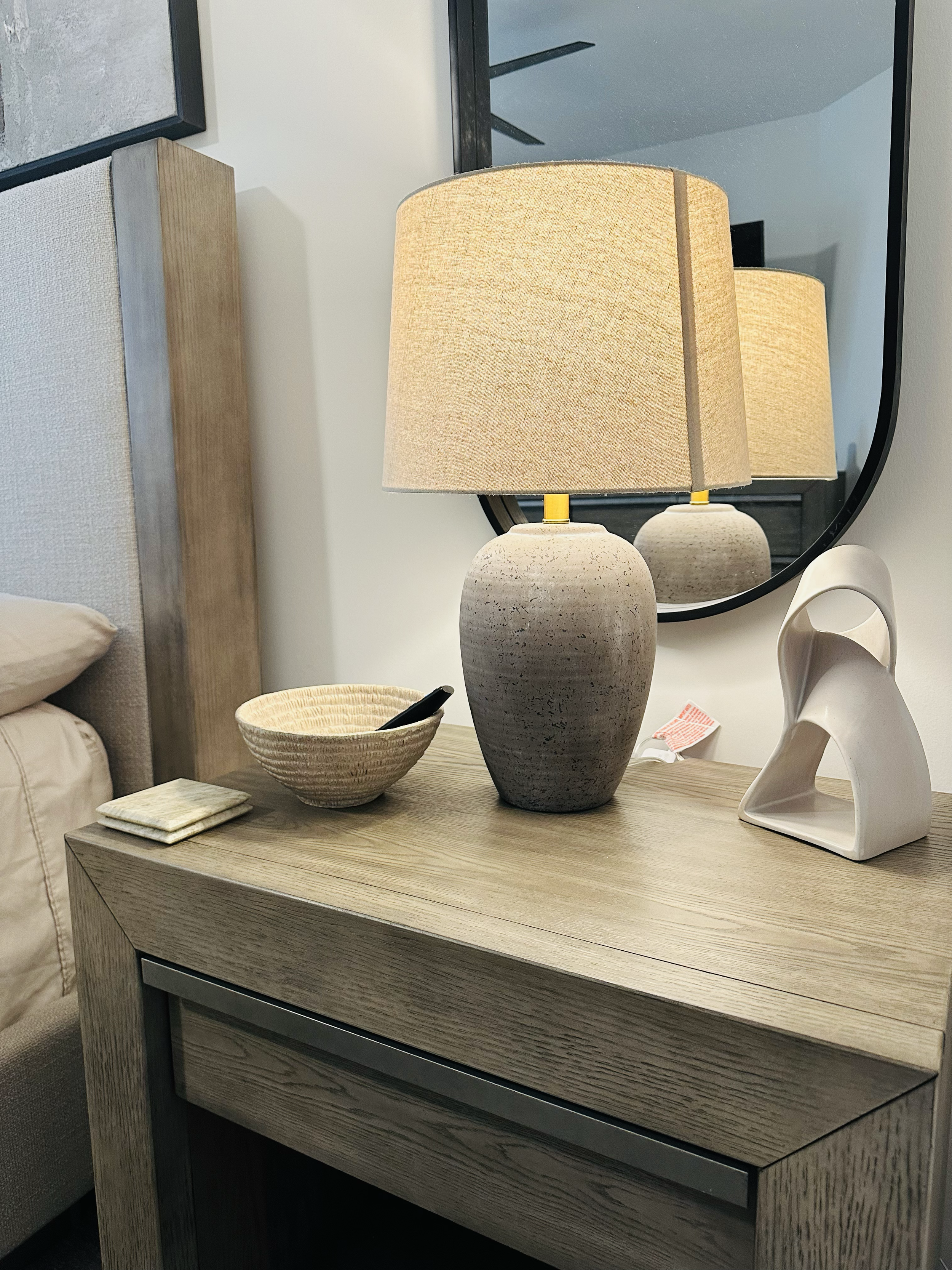 A bedside wooden table with a beige table lamp, a shallow ceramic bowl with a black object inside, a set of small square coasters, and a modern white abstract sculpture. There is a large mirror reflecting the lamp. The background includes a needle-li