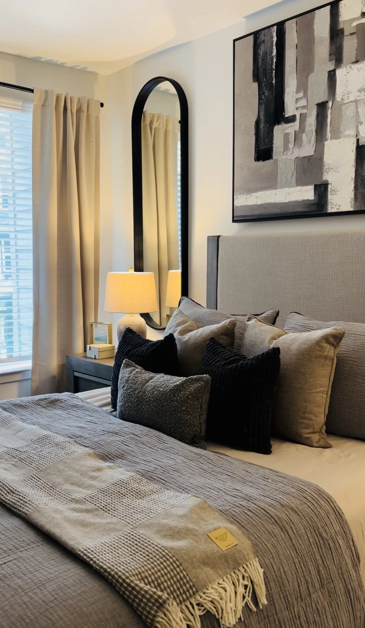 A neatly made bed with a beige headboard, multiple pillows in beige and black, a beige throw blanket, and a yellow tag, next to a nightstand with a lamp, a small framed picture, and some books. There is a large wall mirror and an abstract black and w