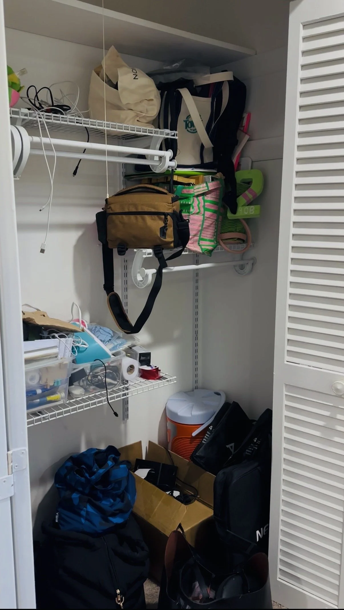 A cluttered closet with shelves holding various bags, boxes, and miscellaneous items, including a brown camera bag, a pink and green striped bag, and a white cooler, along with tangled cords and small containers.