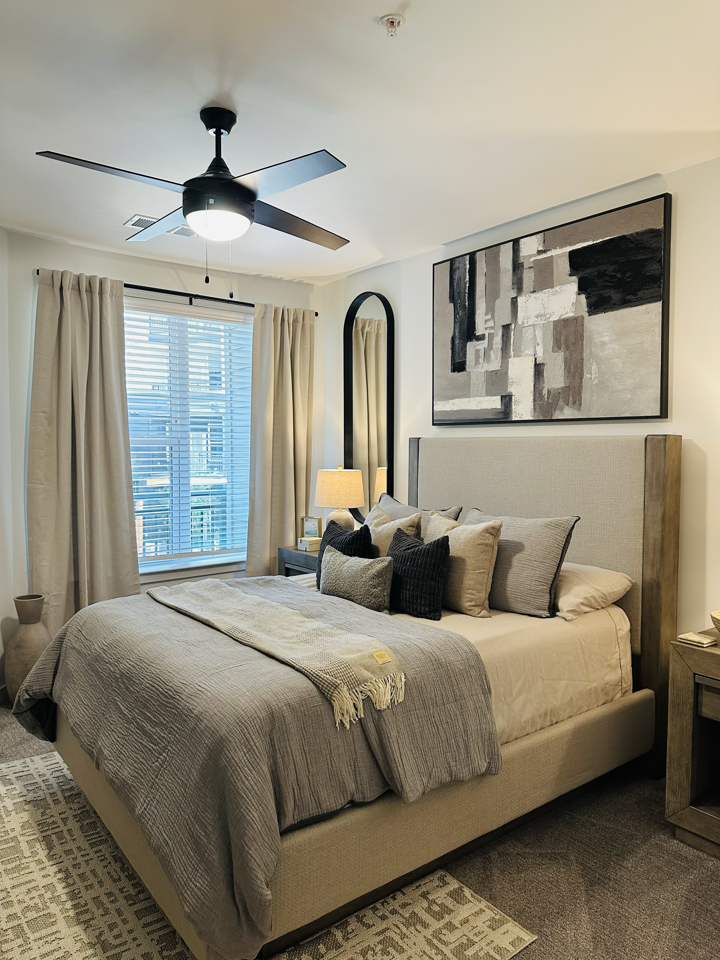 A neatly made bed with beige and gray bedding, multiple pillows, a blanket, and a textured headboard in a modern bedroom. A nightstand with a lamp and framed photo is beside the bed. A large abstract painting hangs above the headboard. The room has b