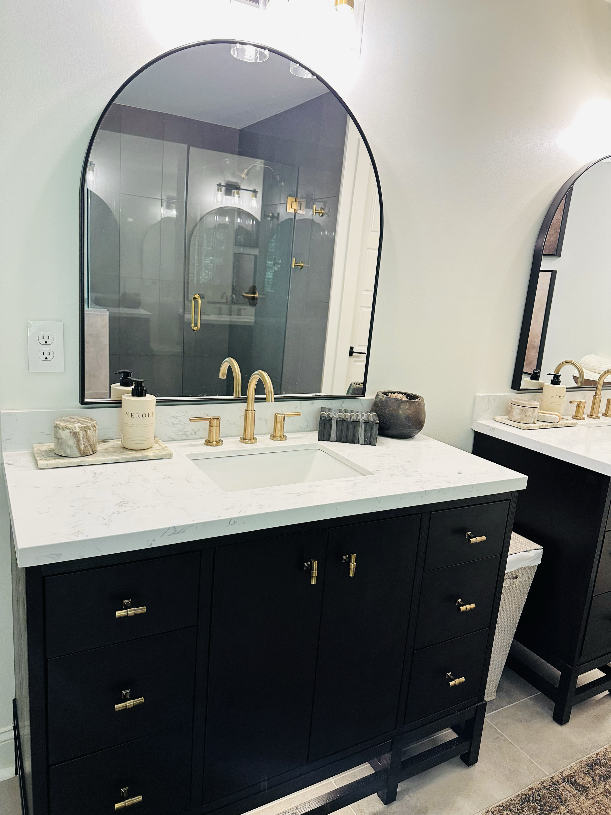 A modern bathroom vanity with a white marble countertop, black cabinetry, and gold hardware. A large arched mirror hangs above the sink, reflecting the shower area with dark tiles and gold fixtures. Decorative items, including a soap dispenser, stone