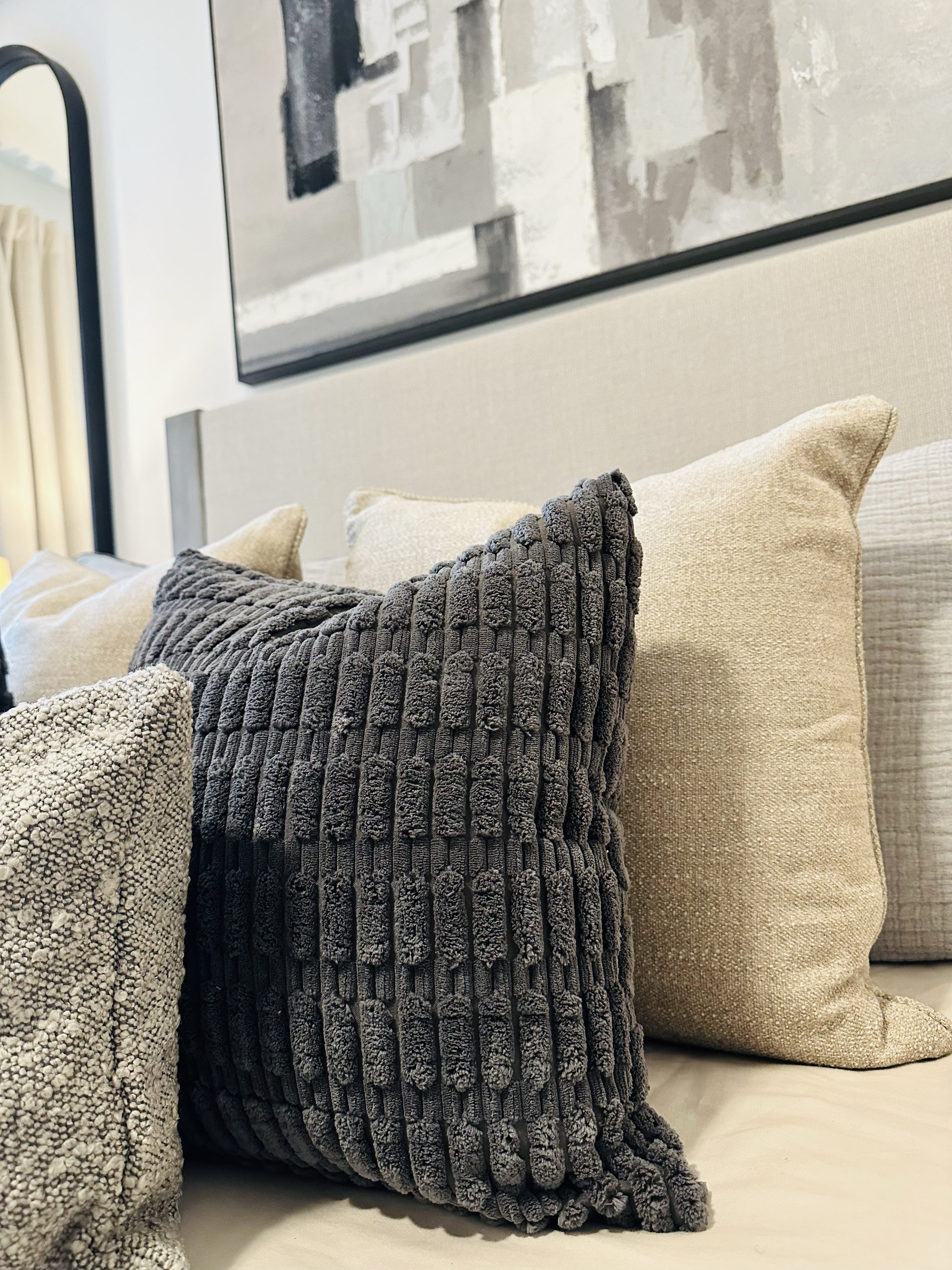 Close-up of beige and dark gray textured throw pillows on a bed, with a beige headboard and abstract artwork above.