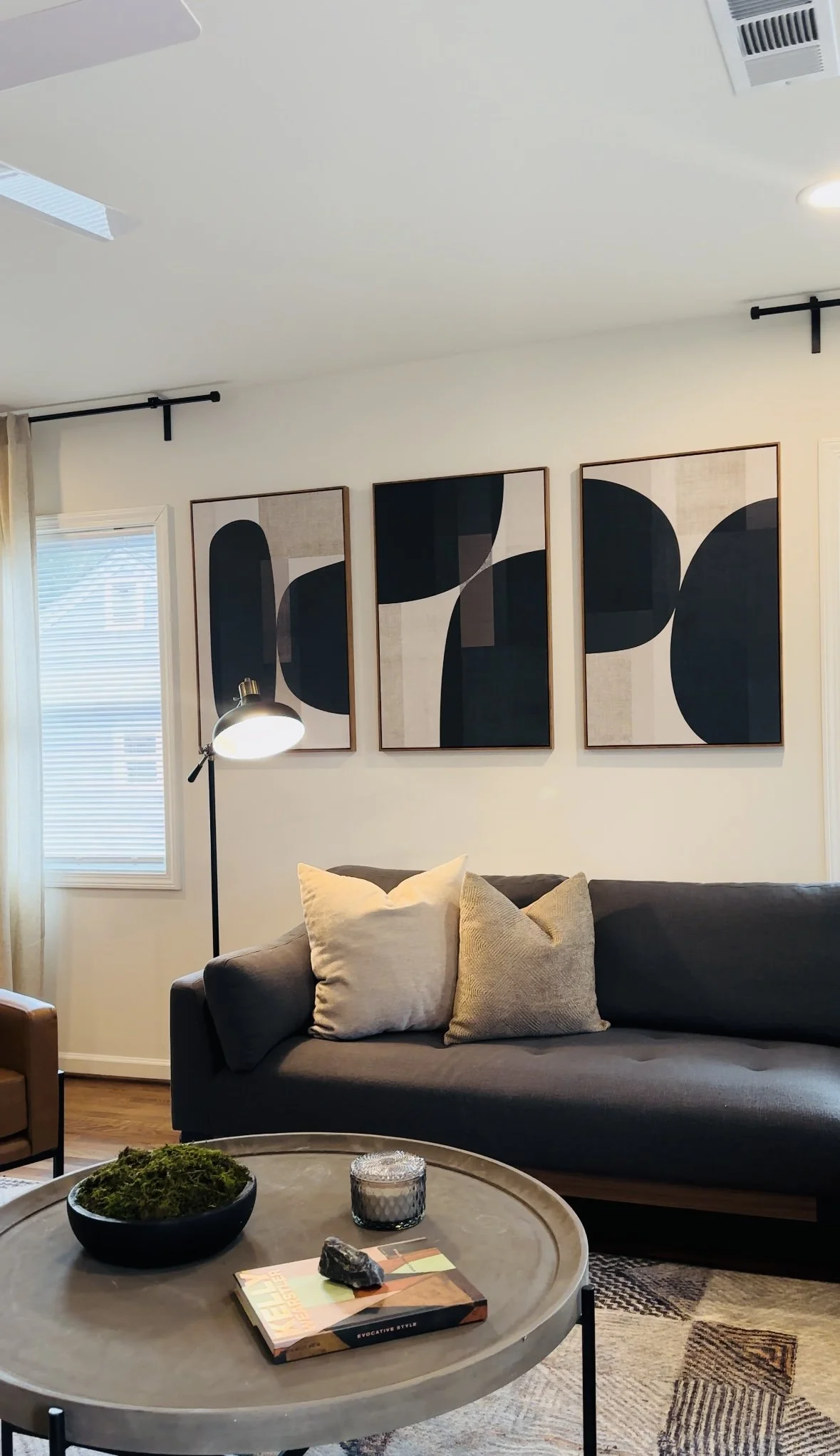 A living room with a dark gray sofa decorated with beige and light brown throw pillows. Above the sofa are three abstract black and beige framed artworks. To the left, there's a window with blinds and beige curtains. In front of the sofa, a round cof