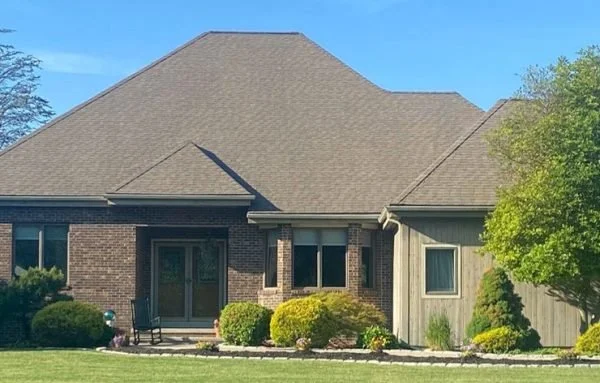 A residential house with a large shingled roof, brick and wooden exterior, surrounded by a well-maintained lawn and bushes.