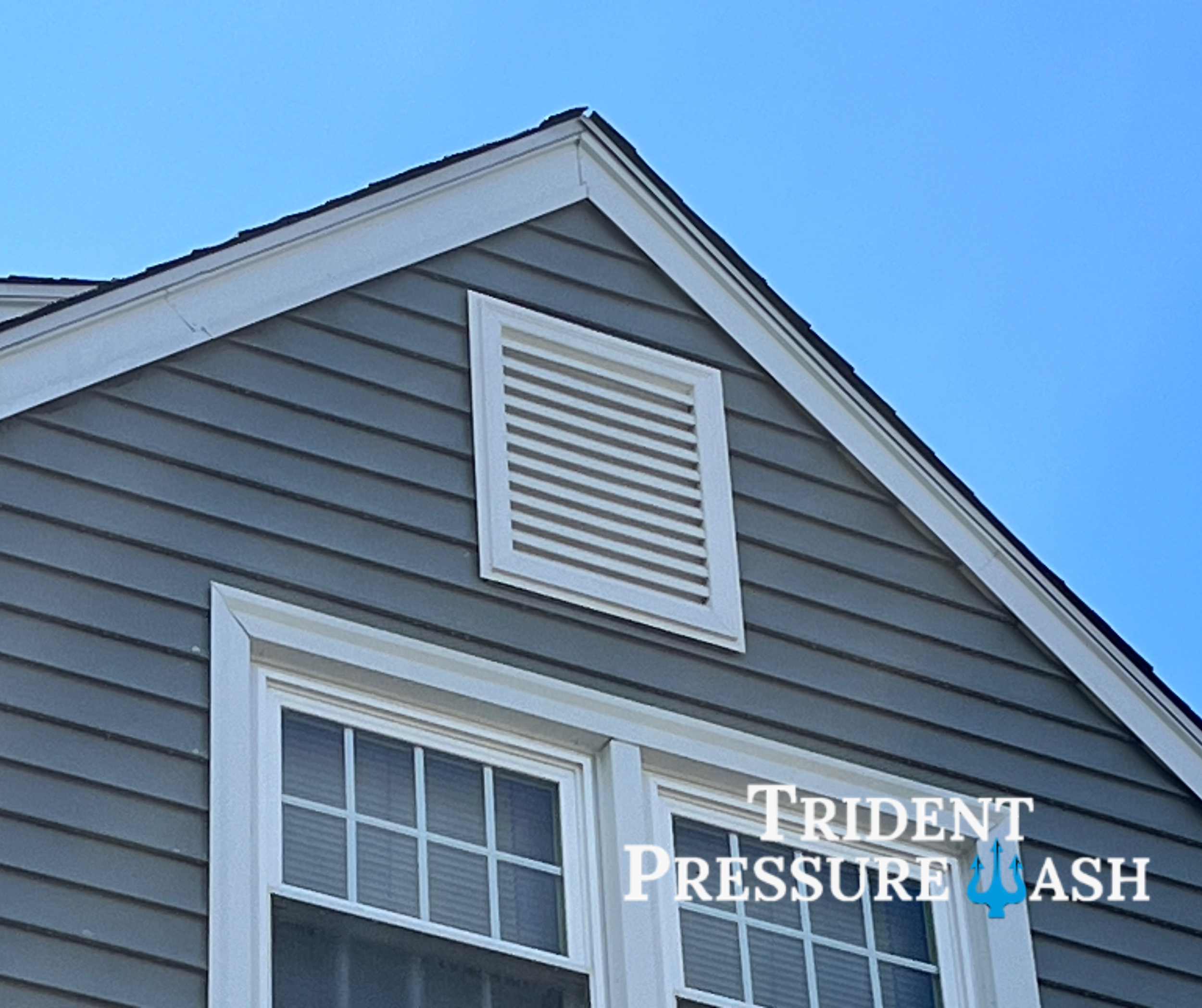 Close-up of the upper part of a gray house with white trim, showing a window and a vent on the gable. There is a logo with a trident and the text 'Trident Pressure Wash' overlaid.