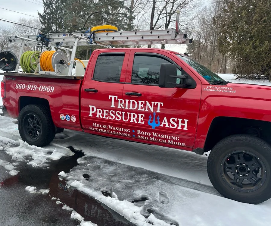 Red pickup truck with logo and contact information for Trident Pressure Wash on the door, parked in snow, carrying ladders, hoses, and cleaning equipment.