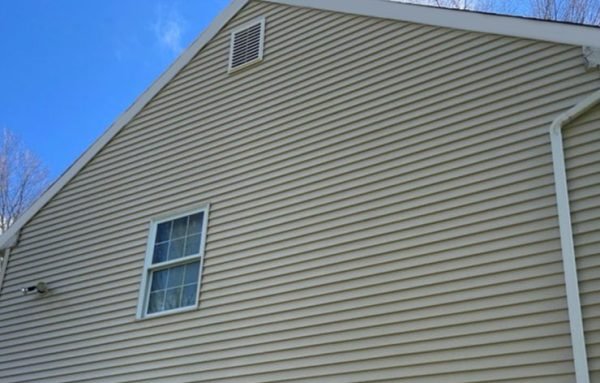 Close-up of the beige vinyl-sided exterior of a house with a window and an attic vent, under a blue sky.