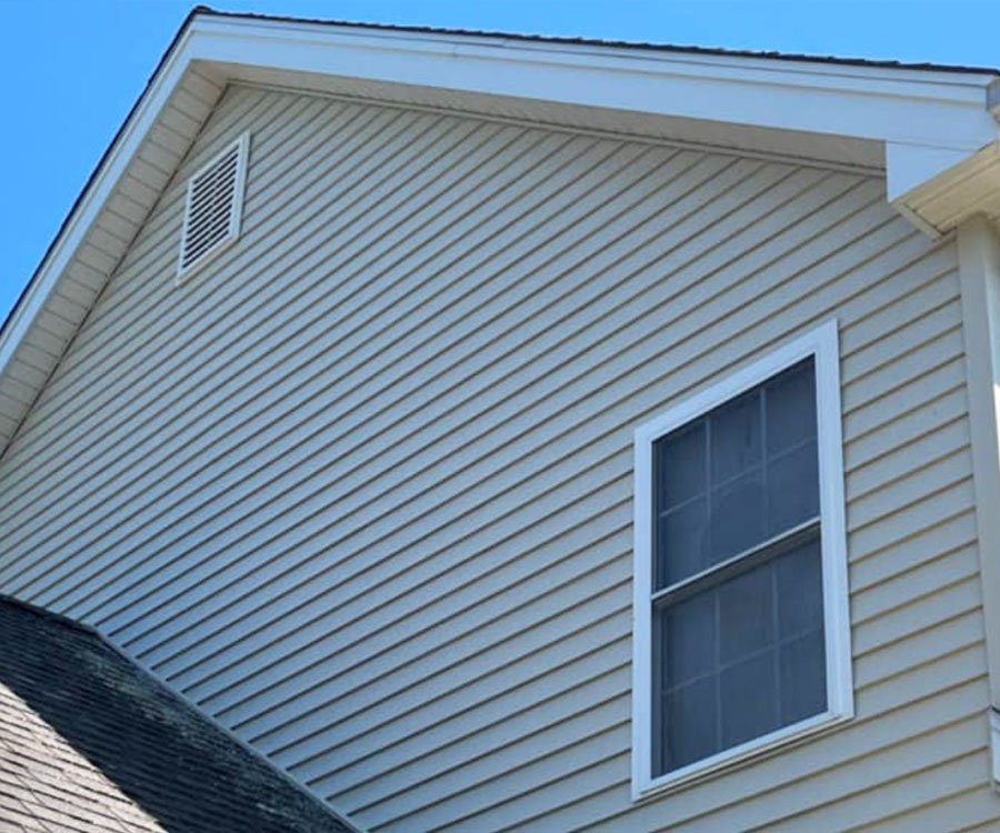 Close-up view of the side of a house with beige vinyl siding, a window, and a small vent near the roof peak.