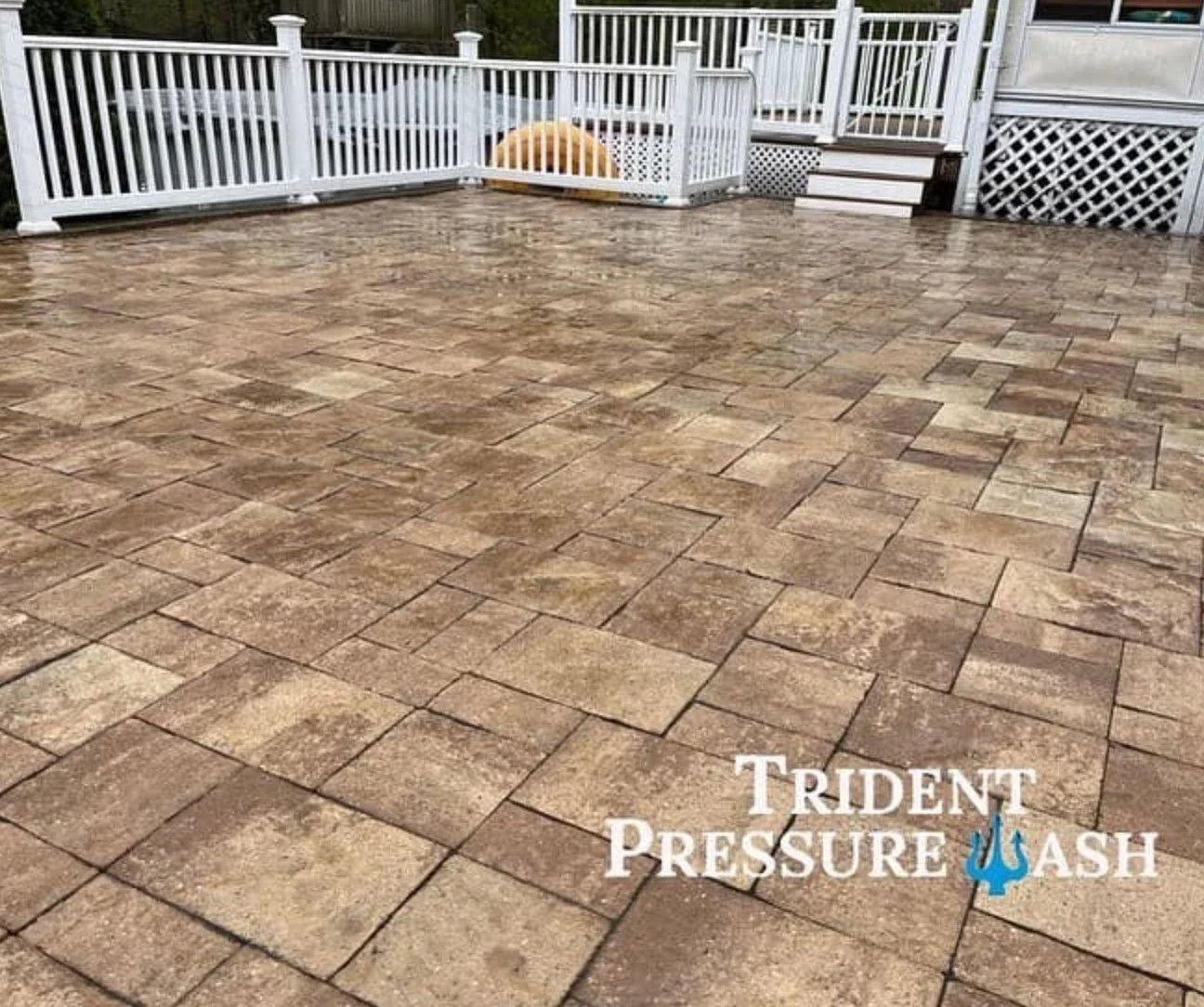 Clean, wet, brown stone patio with white railing and stairs in the background, and a logo that says 'Trident Pressure Wash' in the lower right corner.