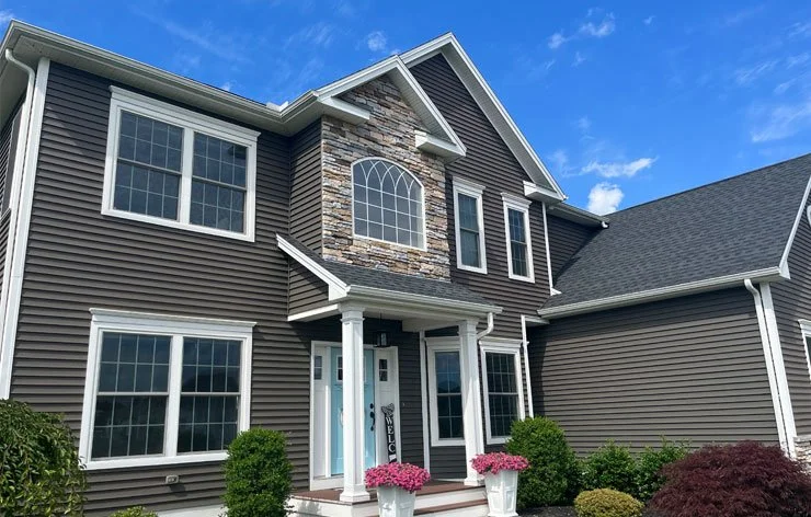 Front view of a two-story house with dark gray siding, white trim, a stone accent wall, and a gabled roof, featuring large windows and a small porch with potted flowers.