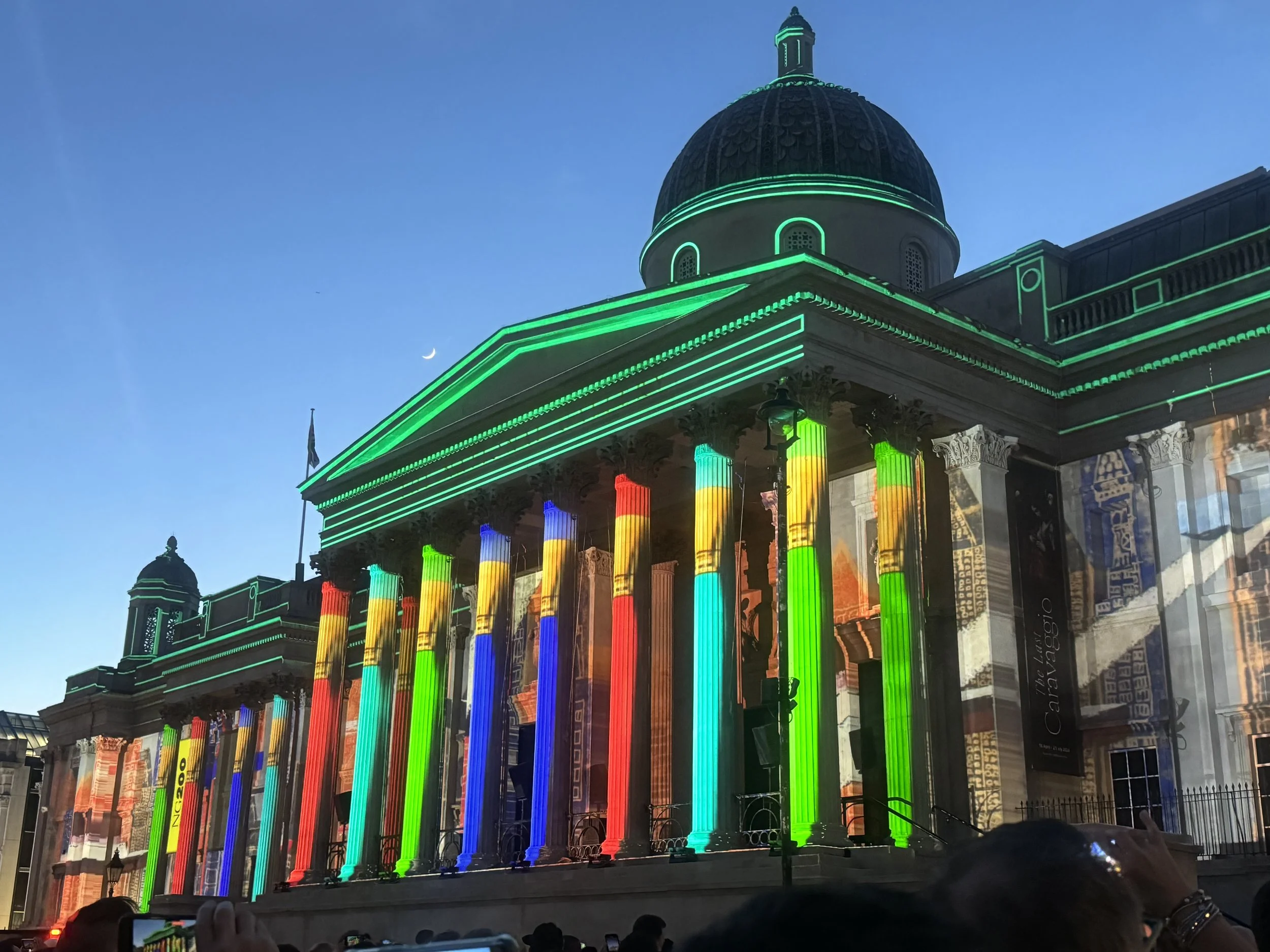 A historic building with a large dome, columns illuminated with rainbow-colored lights, and green neon outline trim, captured during twilight.