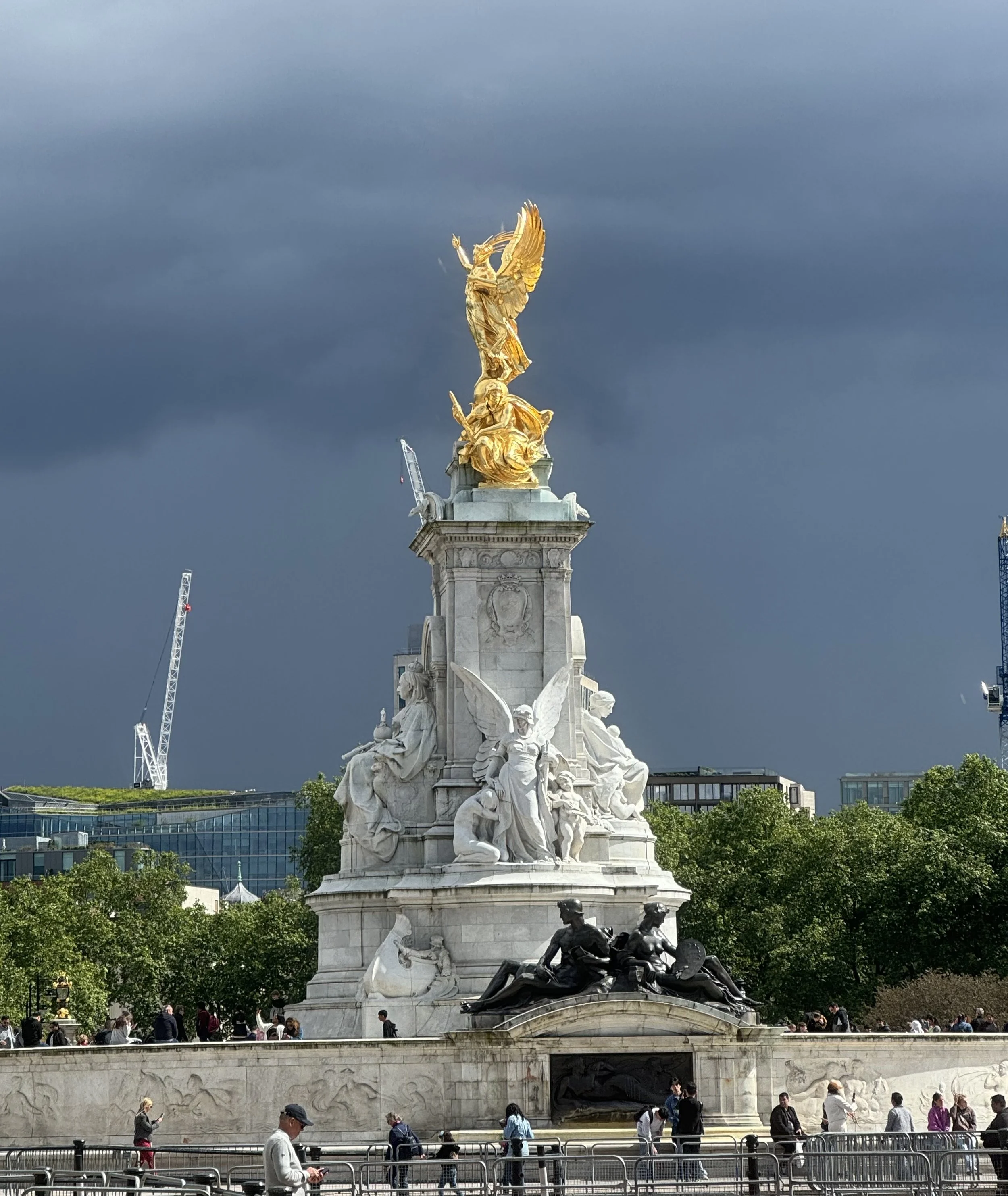 A view of the Victoria Memorial in London with a dark, stormy sky in the background, featuring a white marble and gold statue at the top.