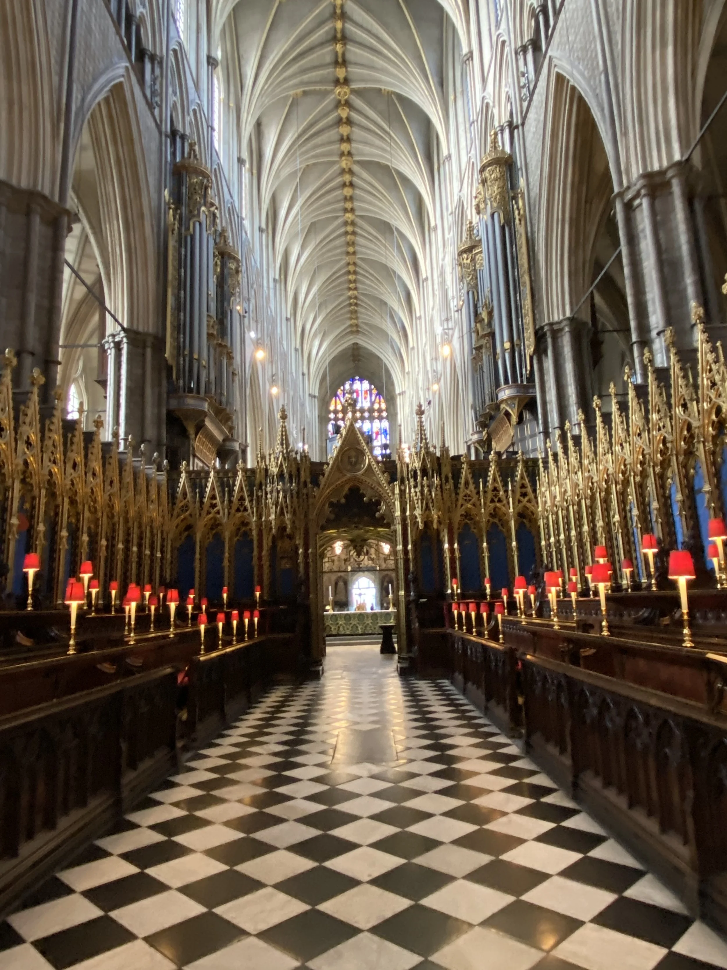 Interior view of a Gothic-style church with high vaulted ceilings, stained glass windows, and ornate woodwork with red lamps along the sides of the worship area. The black and white checkered floor leads to the altar at the far end.