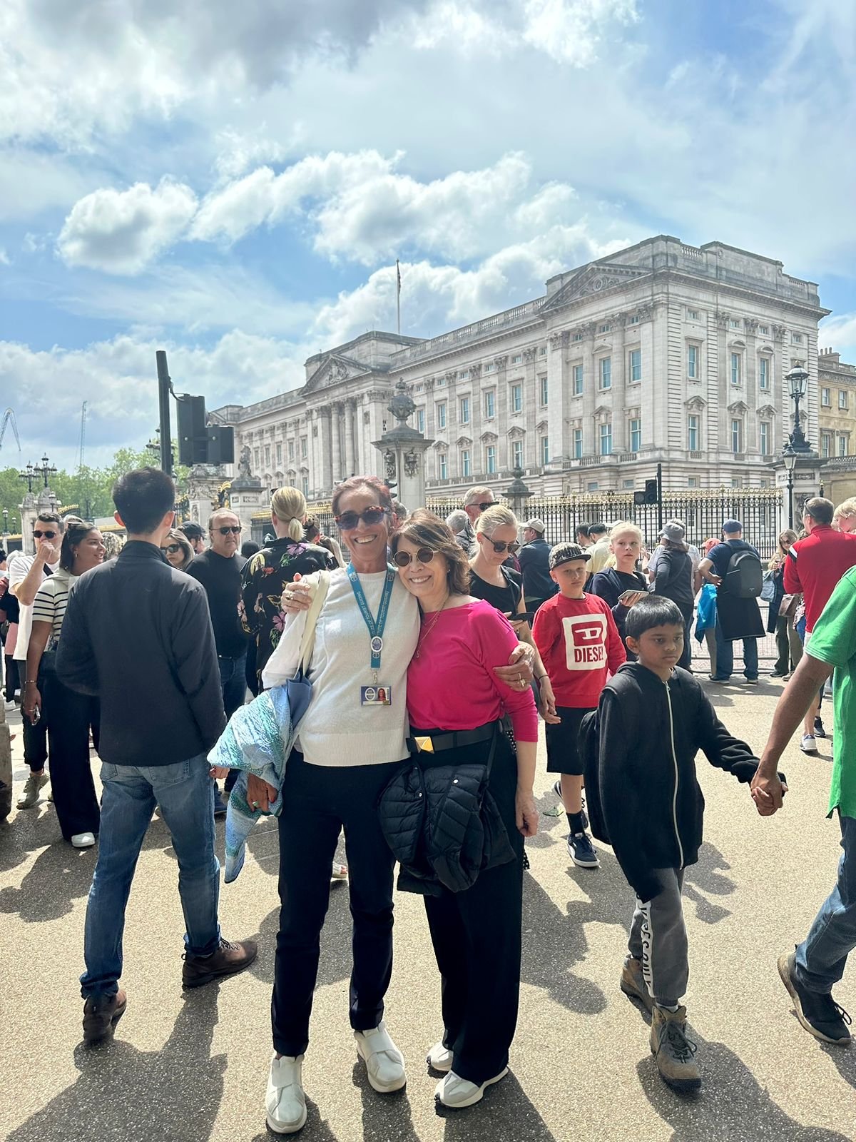 Two women smiling and hugging in front of a crowd of people near Buckingham Palace on a sunny day.