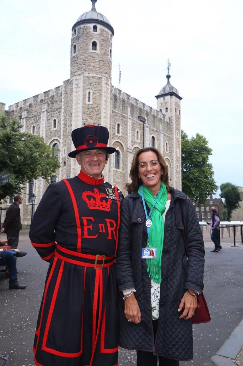 A smiling man dressed as a Beefeater and a smiling woman in a green scarf stand together in front of the Tower of London during daytime.
