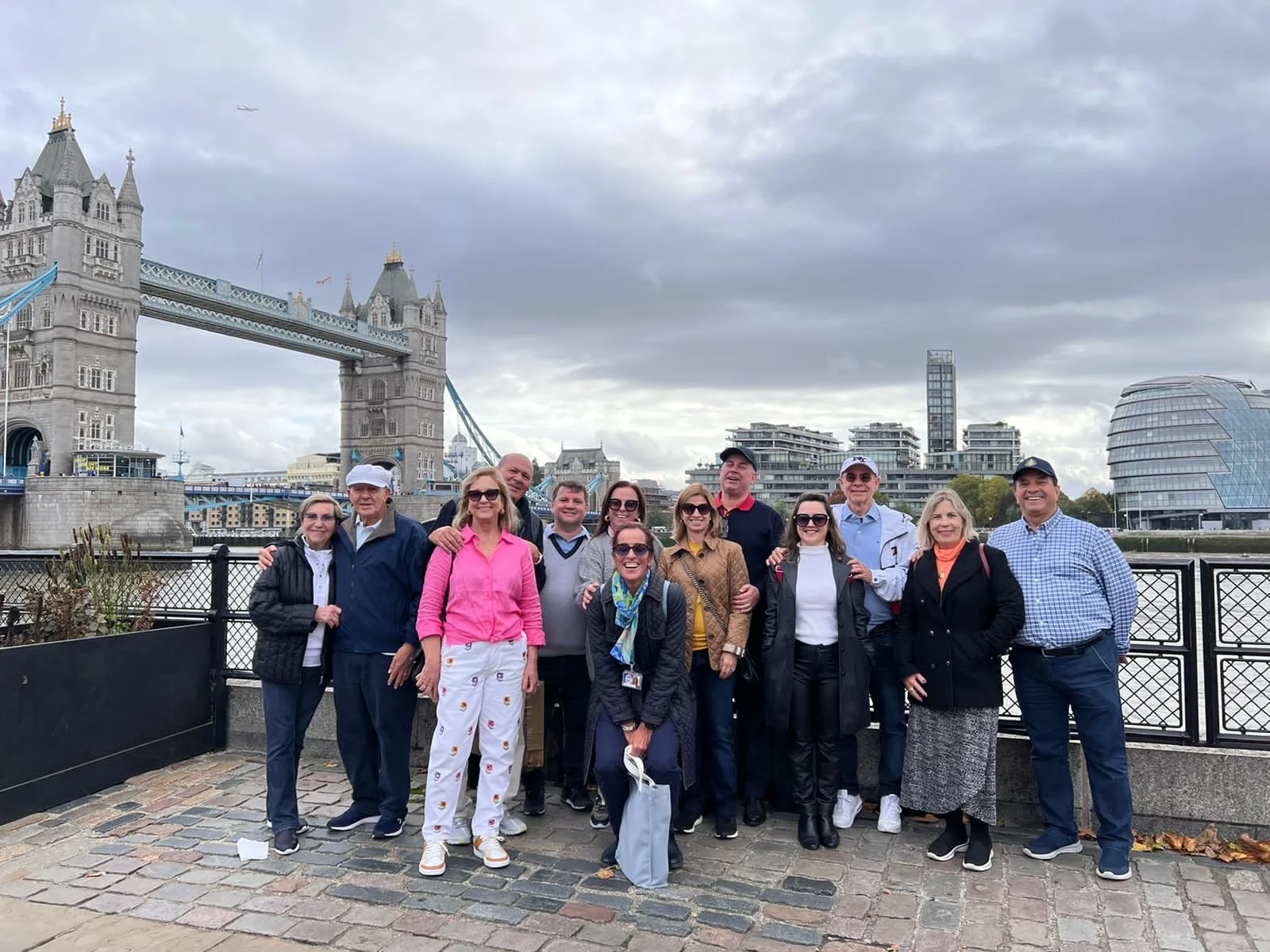 Group of people posing for a photo near Tower Bridge in London, with modern buildings in the background under a cloudy sky.